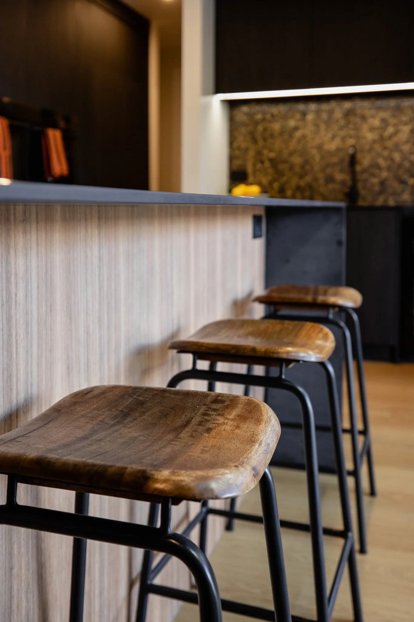 Three wooden bar stools with black metal frames positioned along a light wood bar counter in a modern, dimly lit kitchen or bar area.