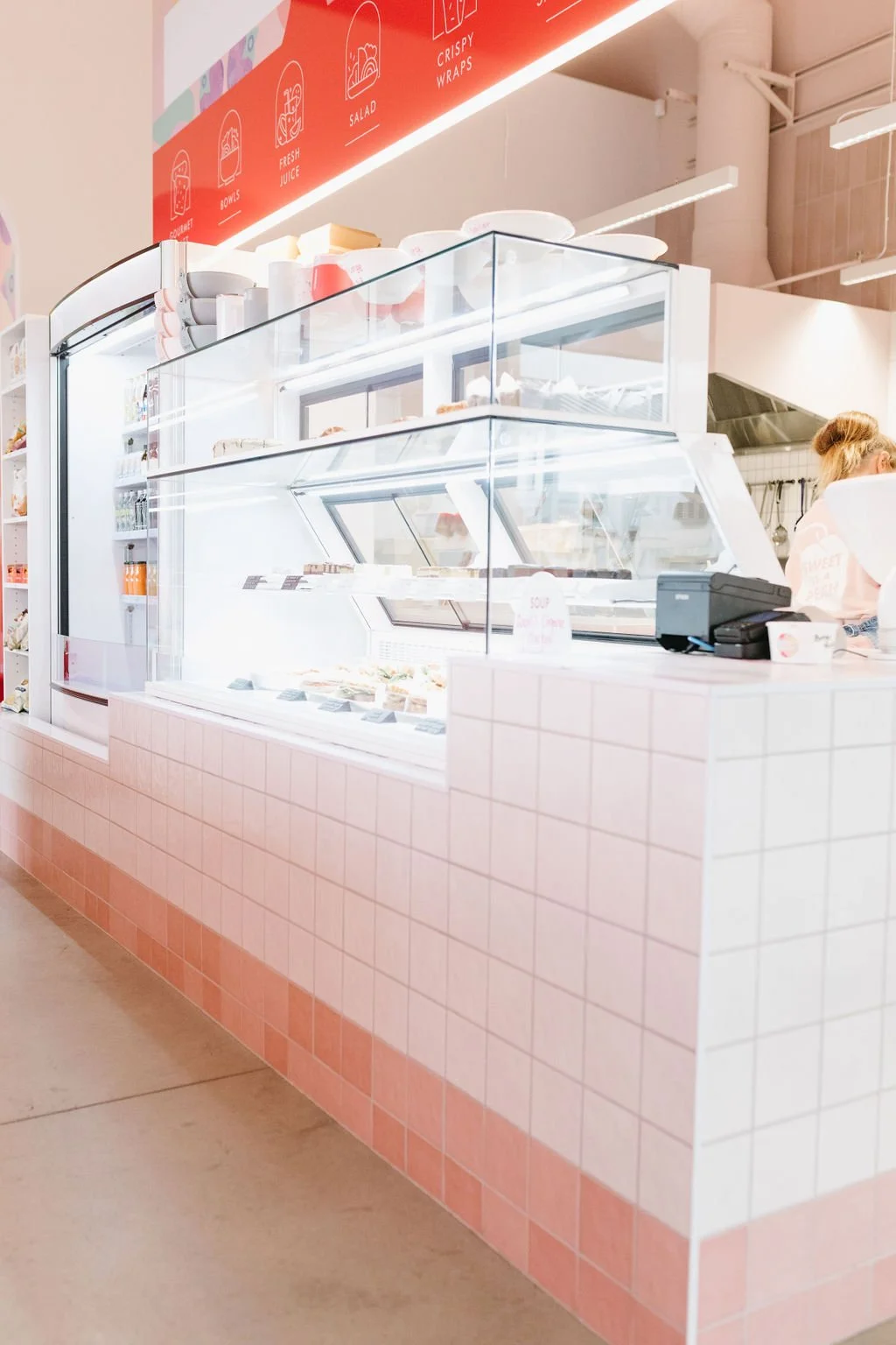 Inside a bright, modern ice cream shop with a glass display case containing various ice cream flavors. The counter is tiled in pink, and a cash register is on the right. A red and white menu sign is visible above, listing items like bowls, fresh juic