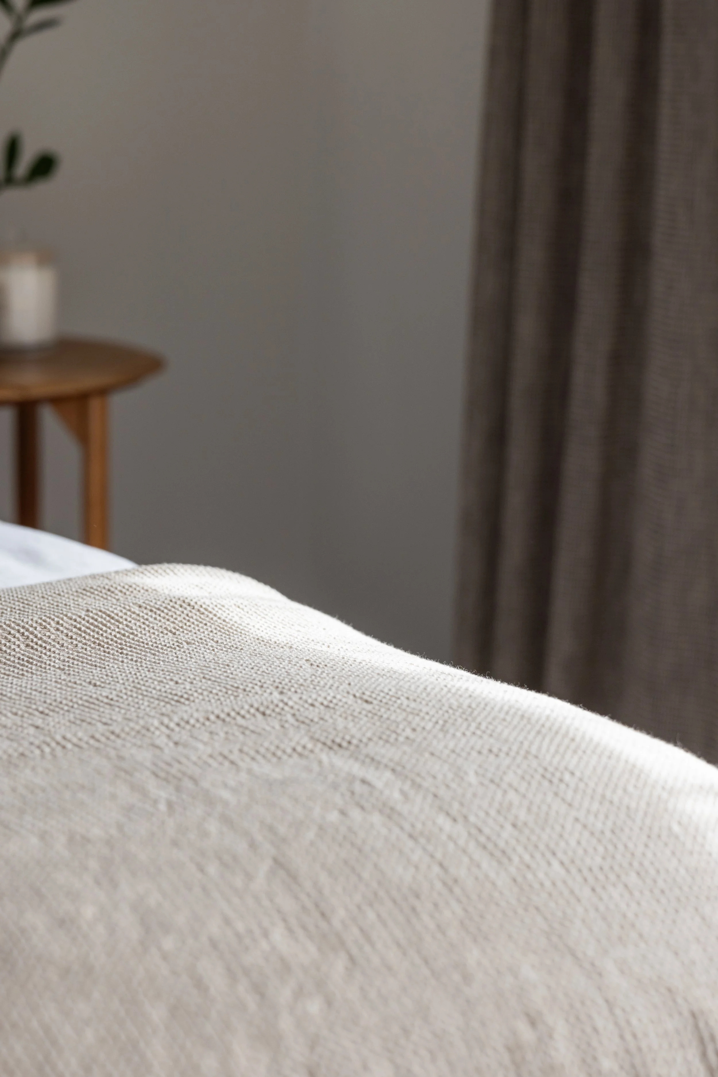 Close-up of a bed with a textured beige bedcover, with a nightstand and a curtain in the background.