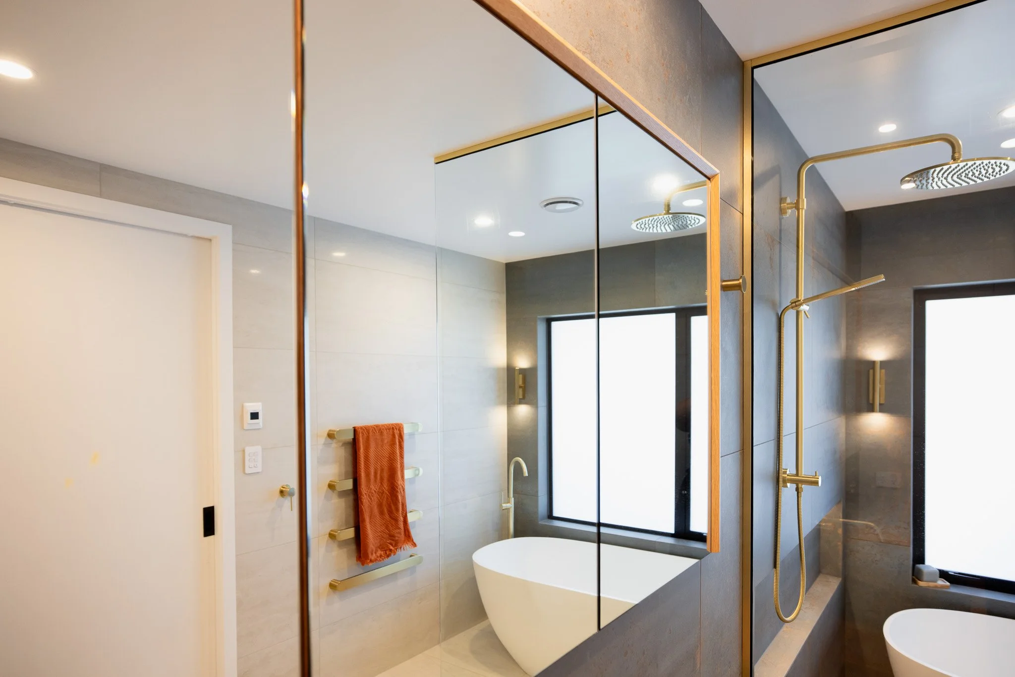 Modern bathroom with a freestanding bathtub, gold shower fixtures, a large shower area with a rain showerhead, a window, and a mirror reflecting part of the room.