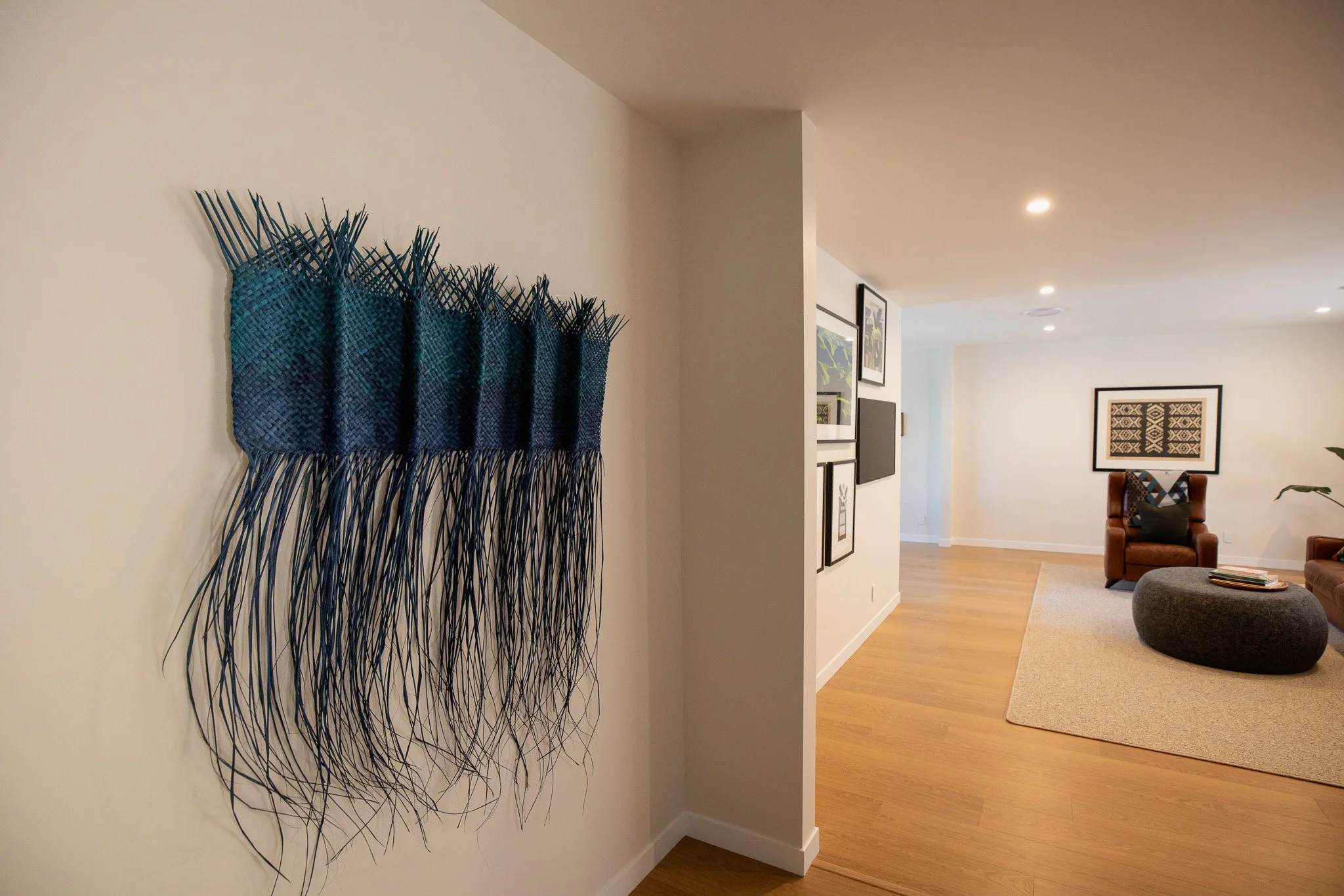 Blue woven wall hanging with fringe on a white wall in a living room with framed artwork, a leather armchair, and a round ottoman.