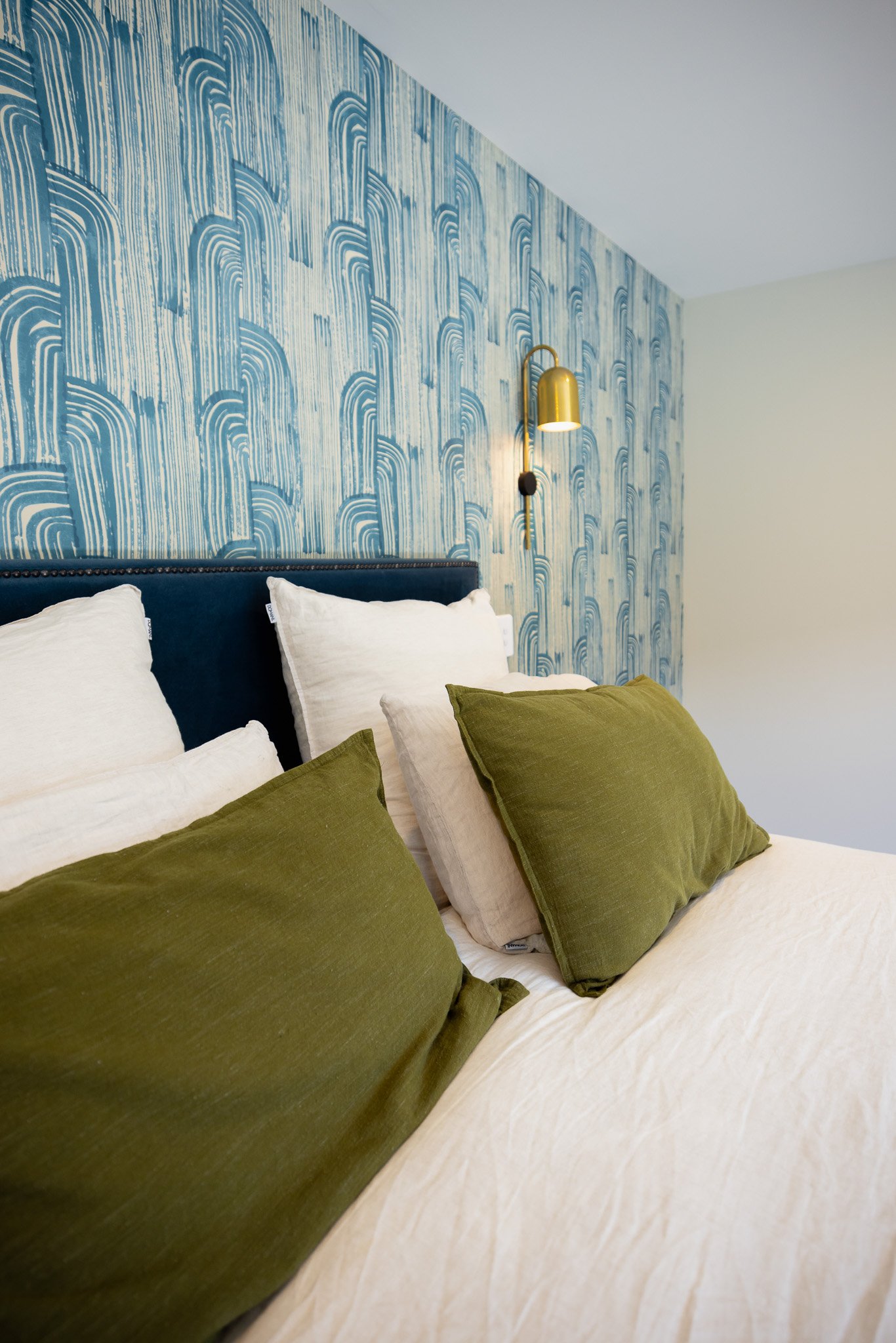 A bedroom with a bed featuring four white pillows and two green pillows, a blue headboard, a patterned blue accent wall, and a gold wall-mounted light fixture.
