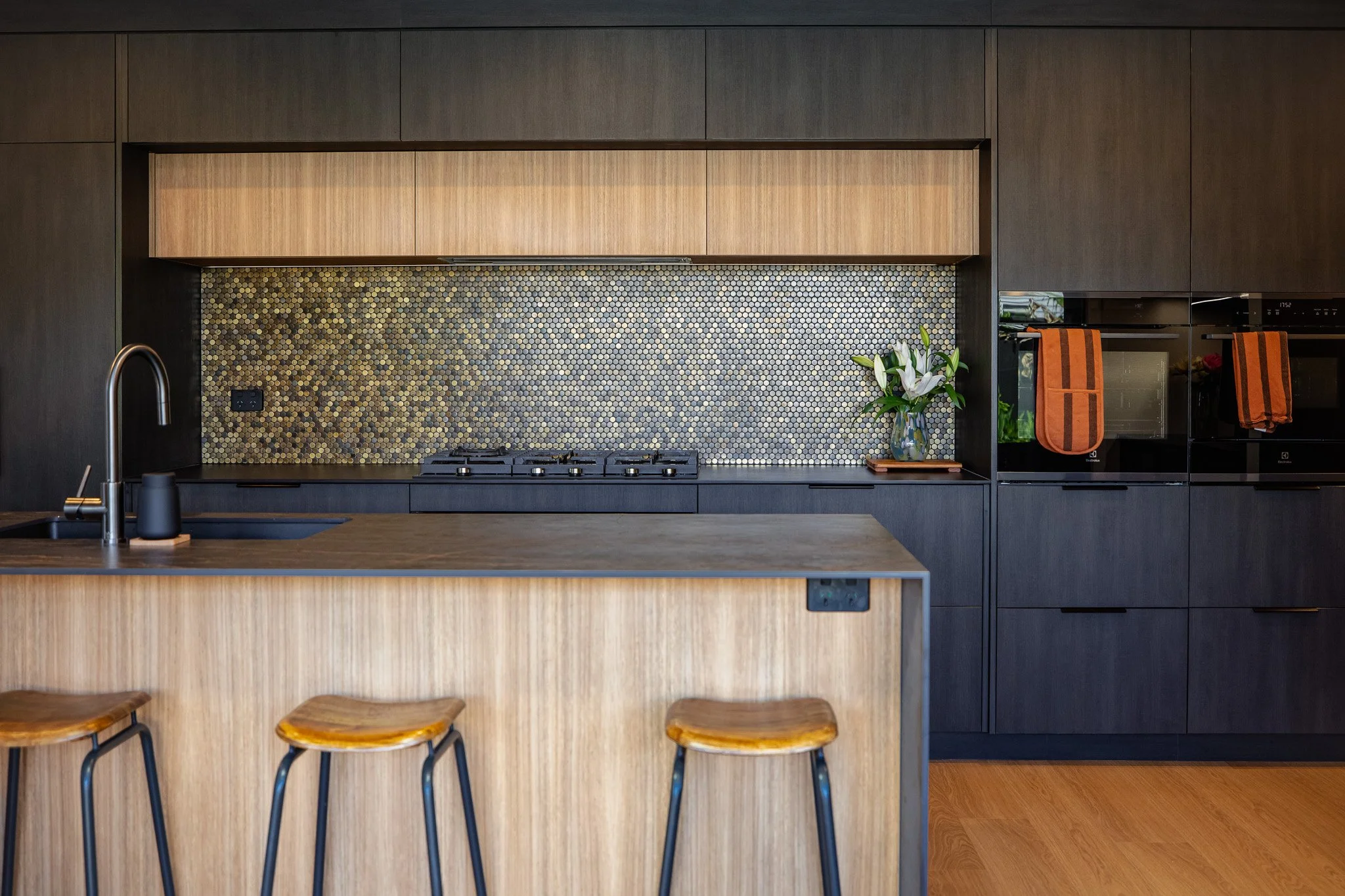 Modern kitchen with dark cabinetry, a light wood island, a honeycomb-pattern backsplash, and a vase of white lilies on the counter.