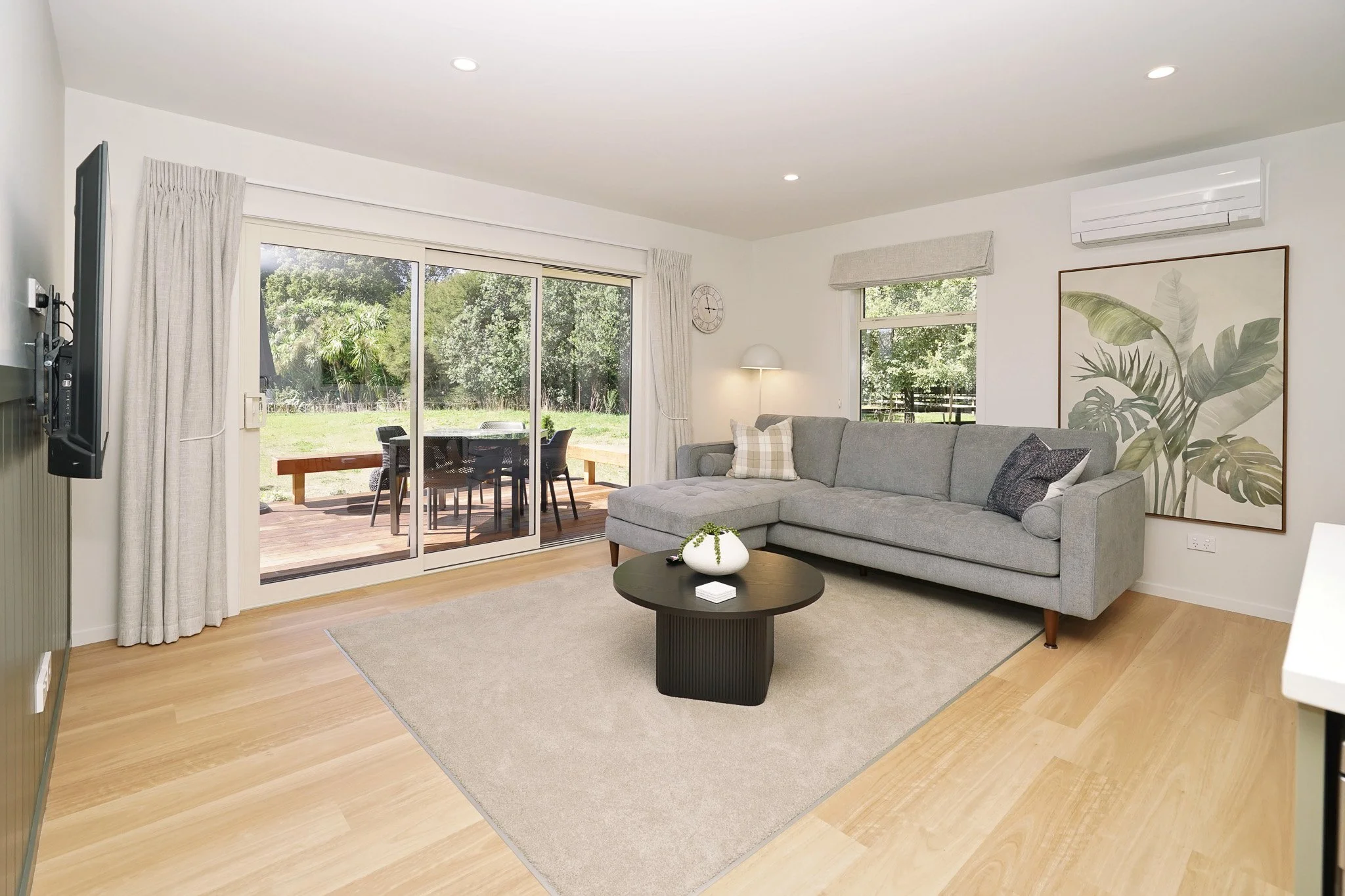 Living room with grey sectional sofa, large window with trees outside, sliding glass door leading to patio with outdoor table and chairs, wall art of large green leaves, beige area rug, wooden floor, side table with white vase, wall clock, air conditioner, and floor lamp.