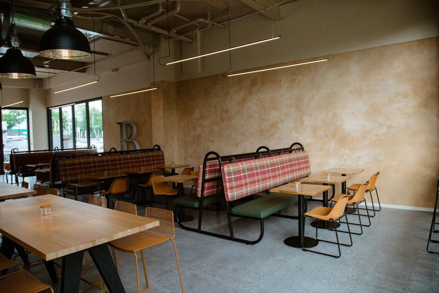 Empty modern cafe with wooden tables, yellow chairs, plaid booth seats, large windows, and industrial-style lighting.