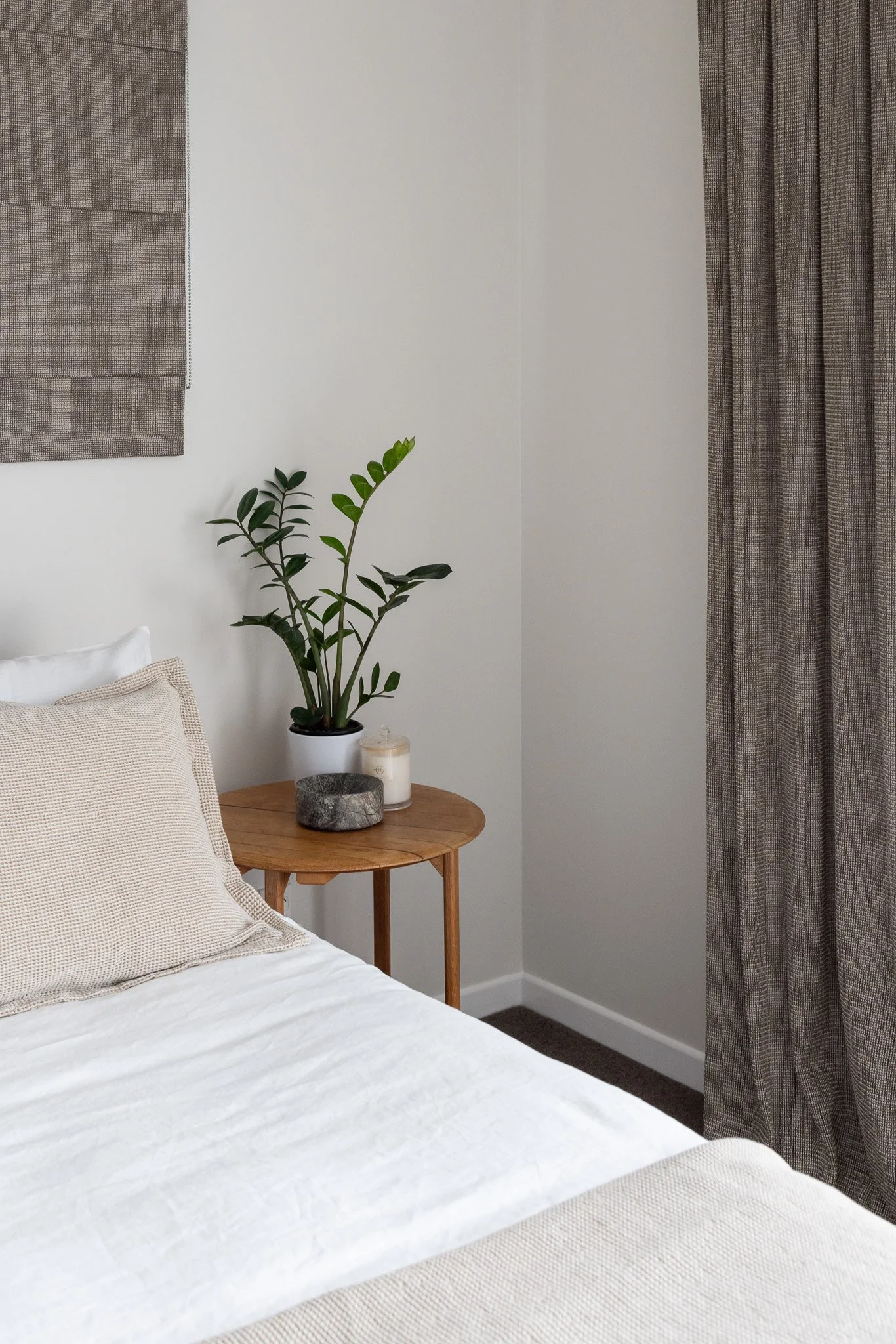 A bedroom corner with a wooden side table holding a potted plant, a candle, and a stone decor piece. Part of a bed with beige and white bedding is visible, with beige cushions. There are grey curtains and a wall hanging behind the bed.