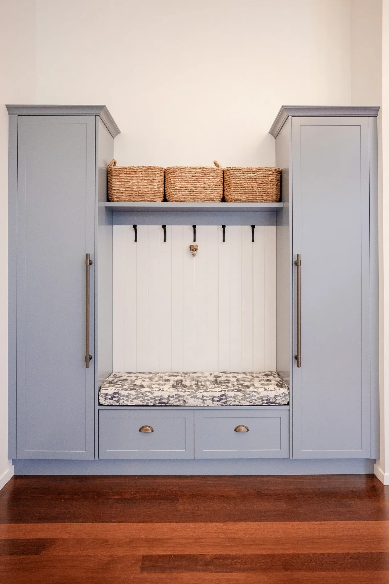 A built-in entryway bench with two tall gray cabinets on each side, a cushion on the seat, overhead shelf with three baskets, and hooks on the wall for hanging items.