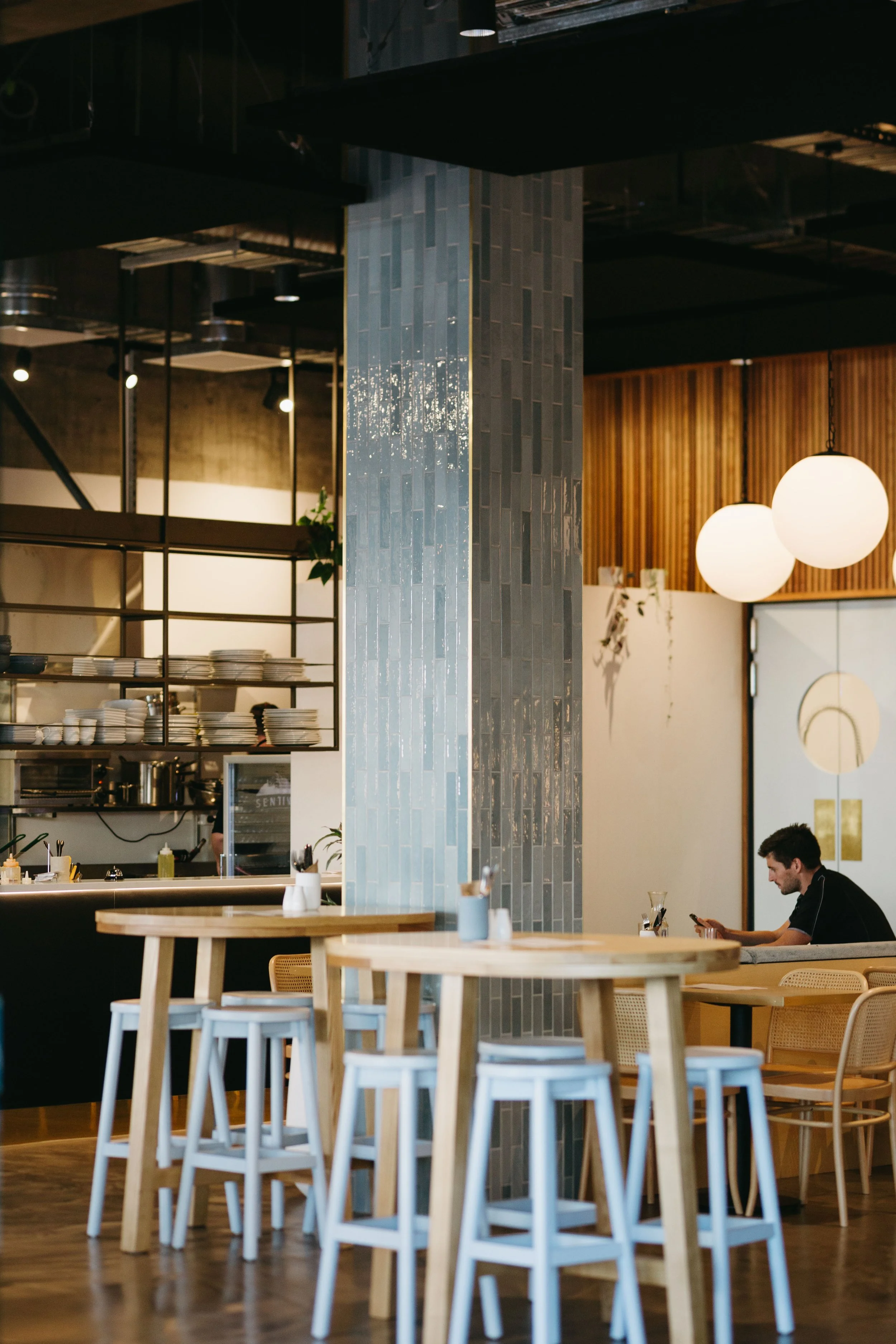 Interior of a modern restaurant with wooden tables and chairs, a decorative tiled pillar, hanging globe lights, and a person sitting at a table using a phone.