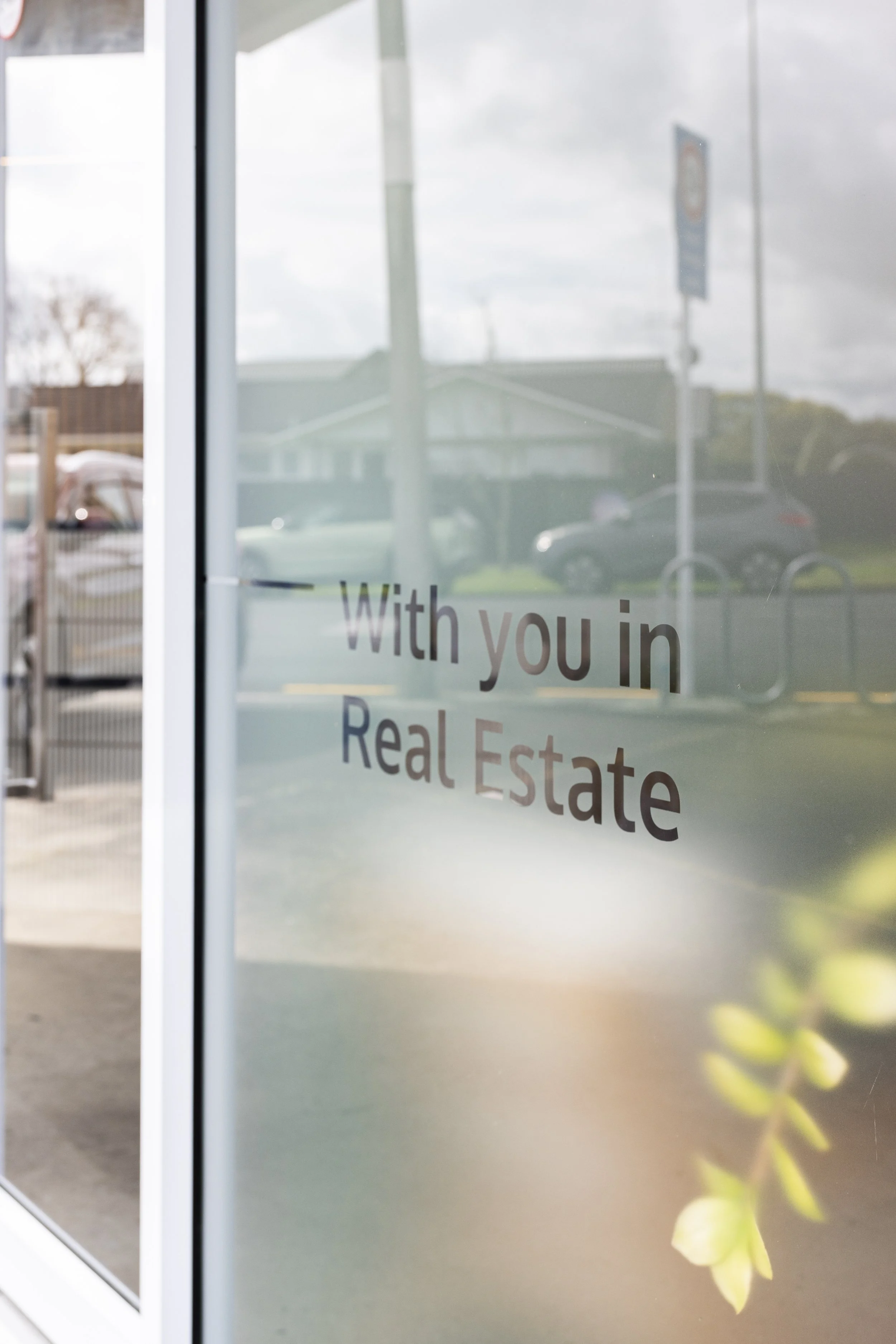 A glass door with the text 'With you in Real Estate' on it, reflecting cars, a house, and clouds outside.