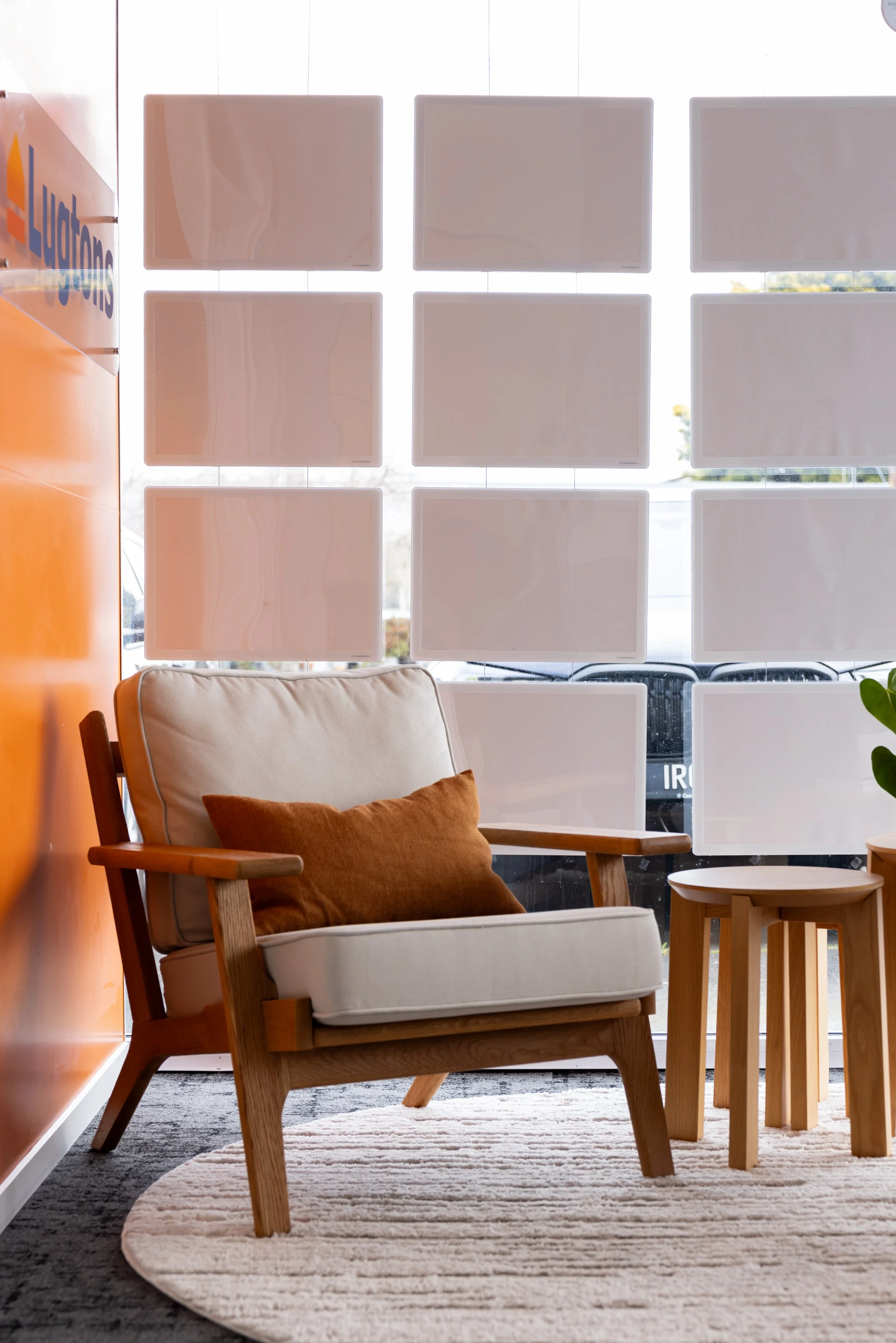 A cozy lounge area with a beige armchair with a brown throw pillow on a round, striped rug, and a small set of wooden nesting tables, in front of a large window with nine white square window panels.