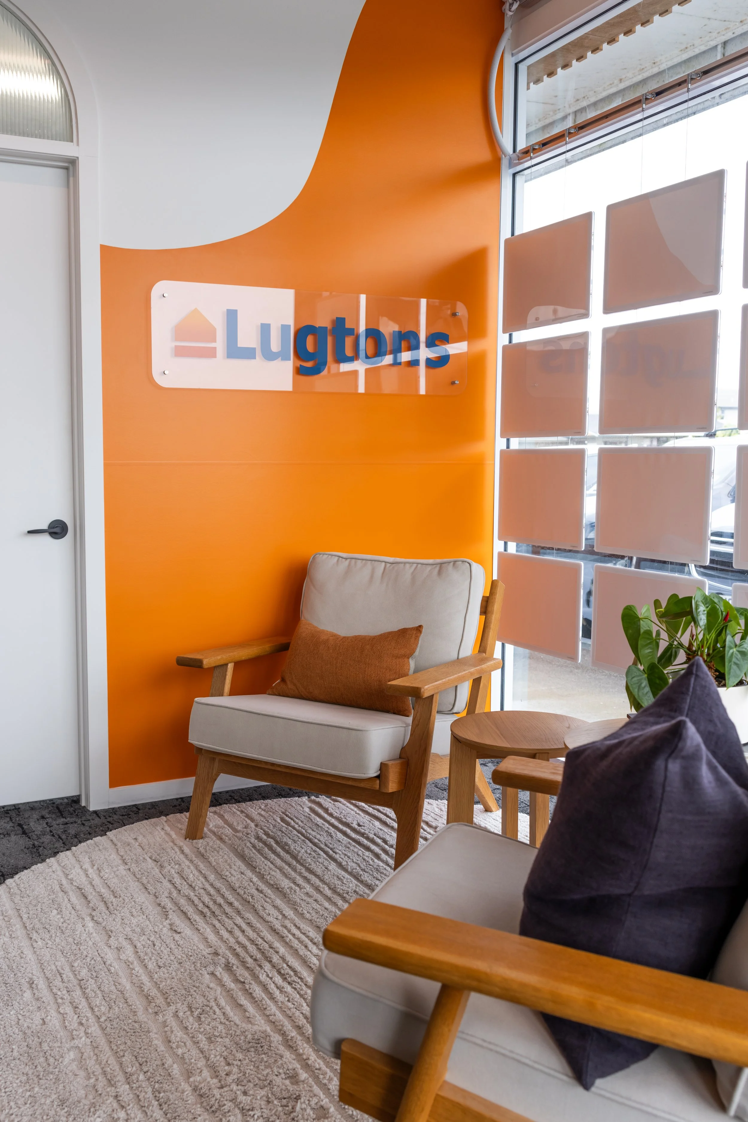 Interior of an office waiting area with orange walls, a sign reading 'Lugtons,' a beige armchair with wooden arms and a brown pillow, a beige sofa with a dark pillow, a small round wooden table, a potted plant, and a window with white cloud-shaped pa