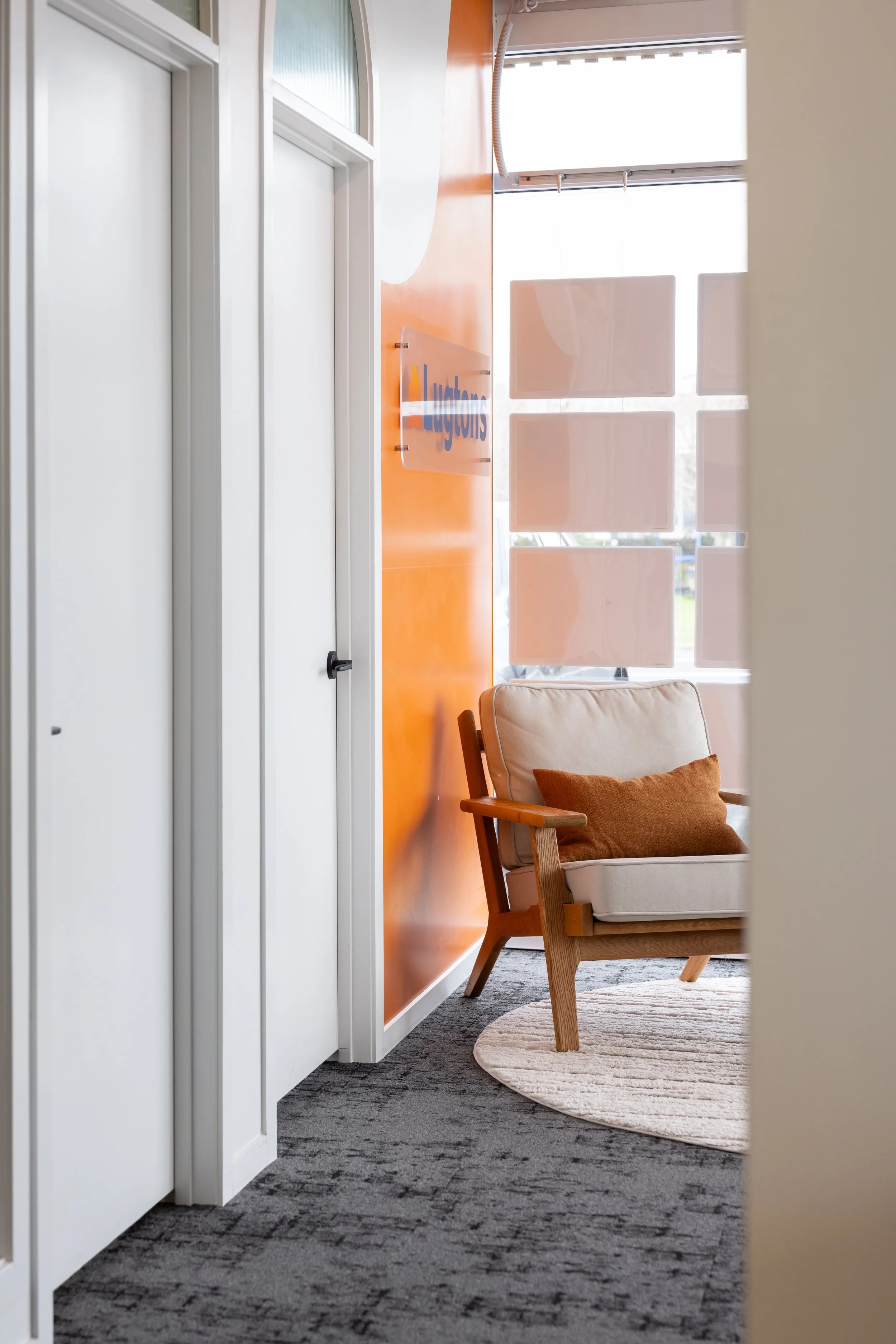 A view of an office waiting area with a beige couch and orange throw pillow, a circular rug, an orange accent wall with a sign, and a window with frosted glass panels.