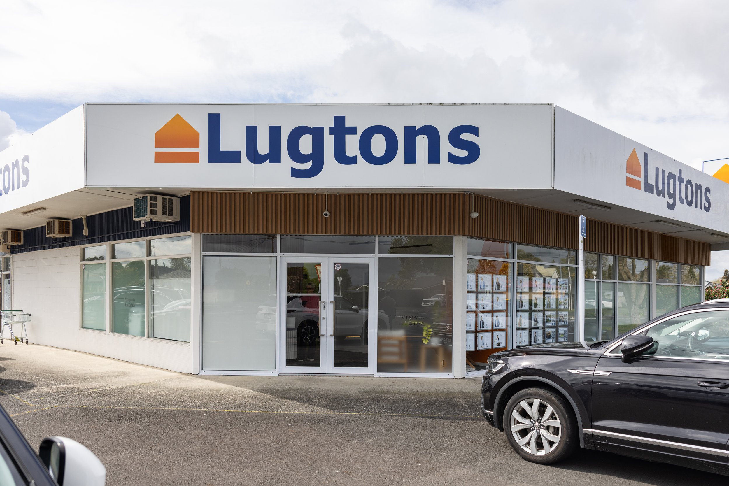 Exterior of a retail store with a sign reading 'Lugtons' and a door with display windows showing posters or images outside. There are cars parked in front of the store.