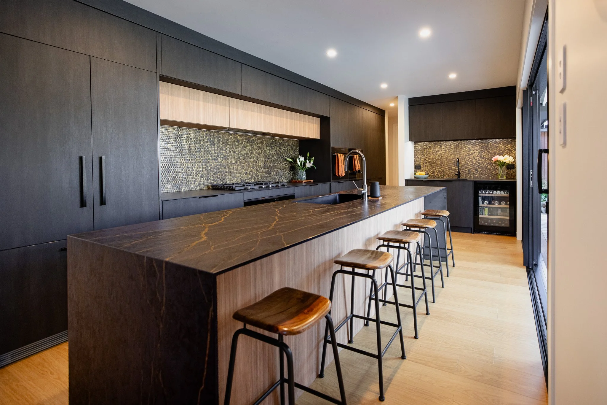 Modern kitchen with dark cabinets, marble island with stools, mosaic backsplash, wine fridge, and minimalist decor.