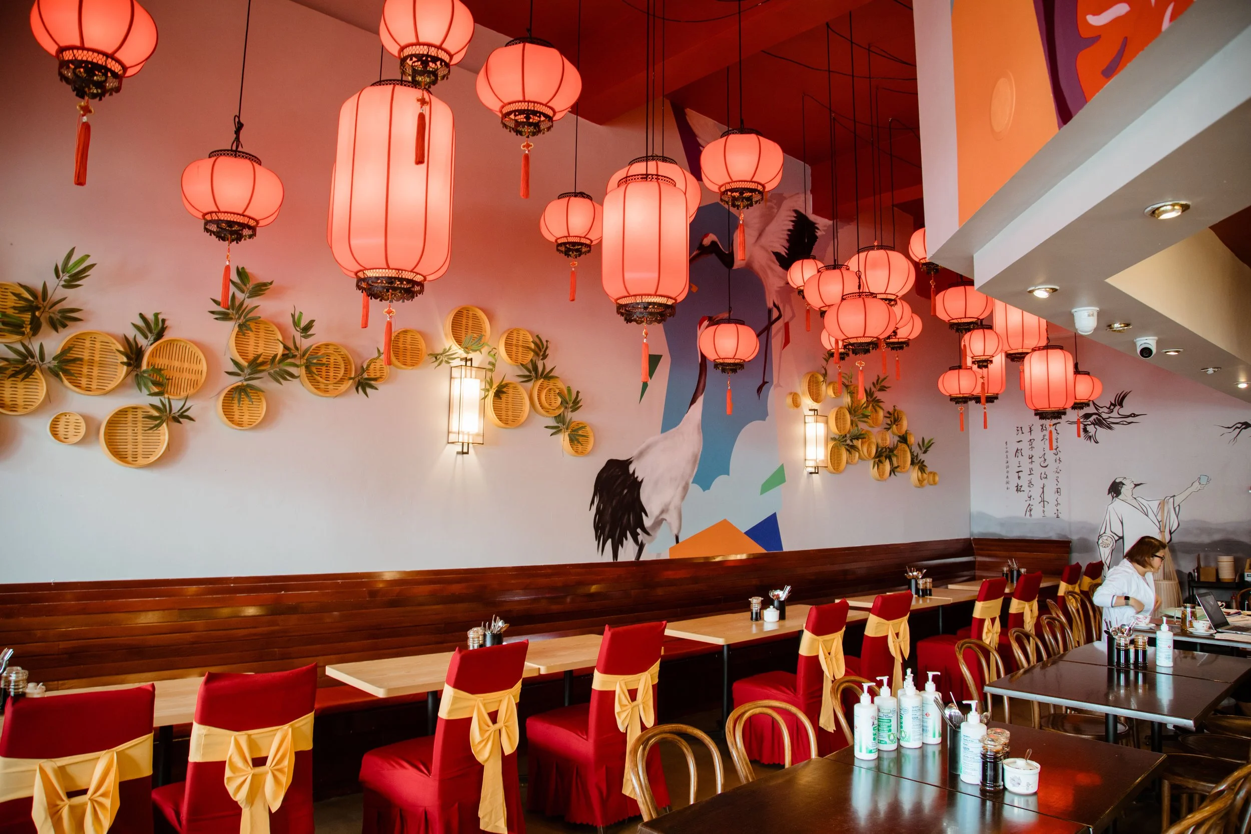 Interior of an Asian-themed restaurant with red hanging lanterns, red chairs with yellow bows, and wall decorations including a crane mural and traditional art.