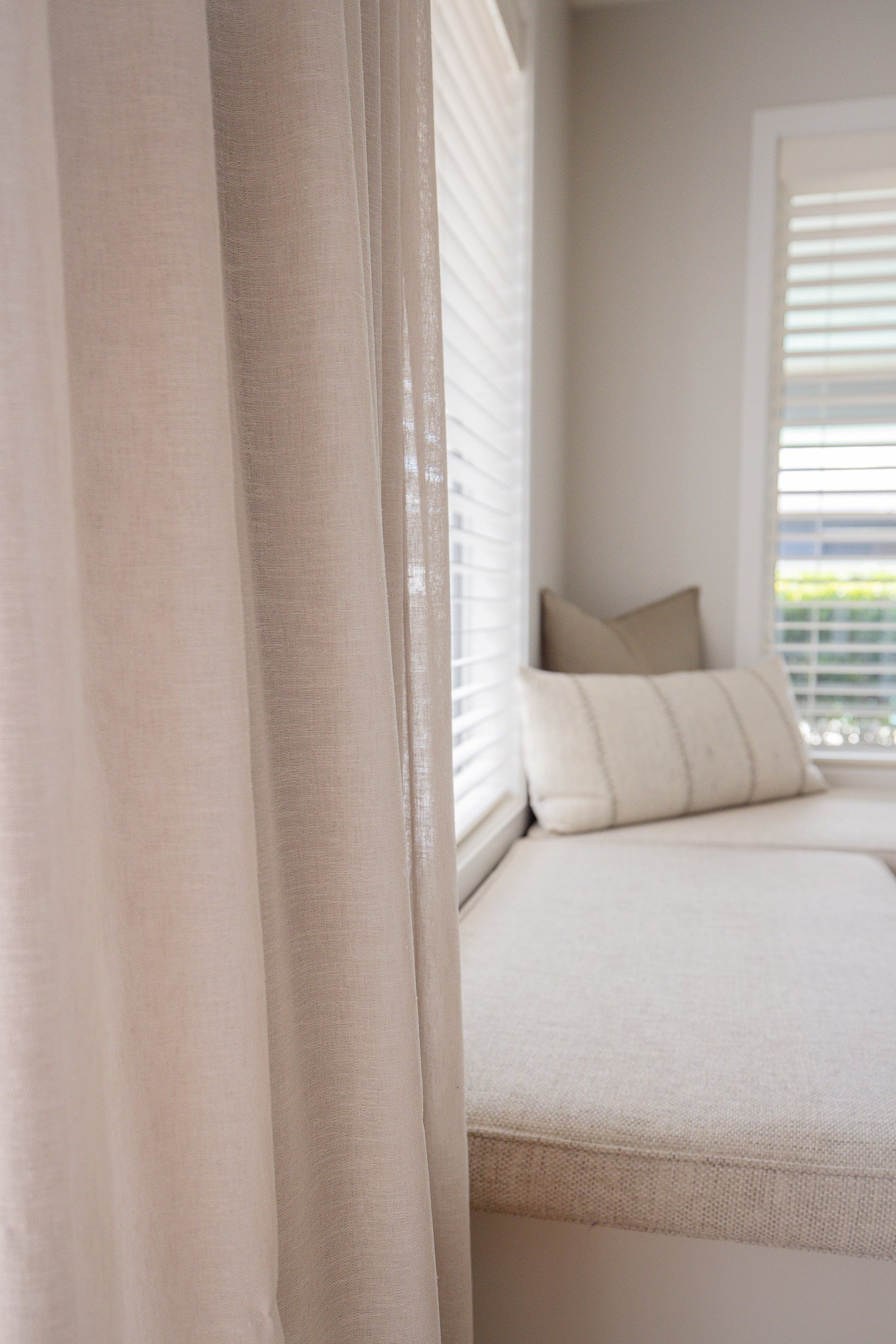 Close-up of beige curtains with a window seat and pillows in the background.