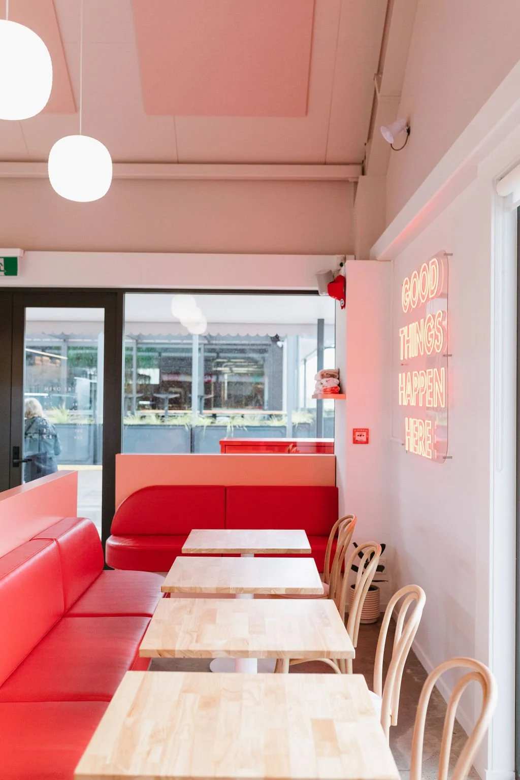 Interior of a modern cafe with red bench seating, light wooden tables, and beige chairs. There is a pink neon sign on the wall that reads 'Good Things Happen Here' and large glass windows showing an outdoor seating area.