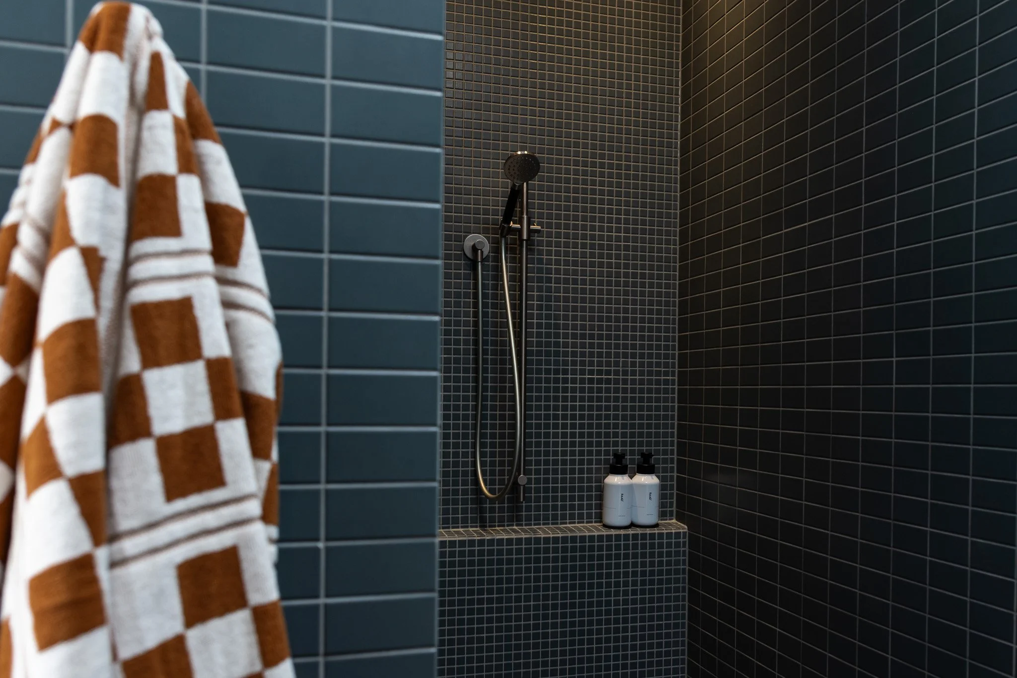 A shower area with dark blue and black tiled walls, a black showerhead, a handheld shower, and two bottles of toiletries on a ledge.
