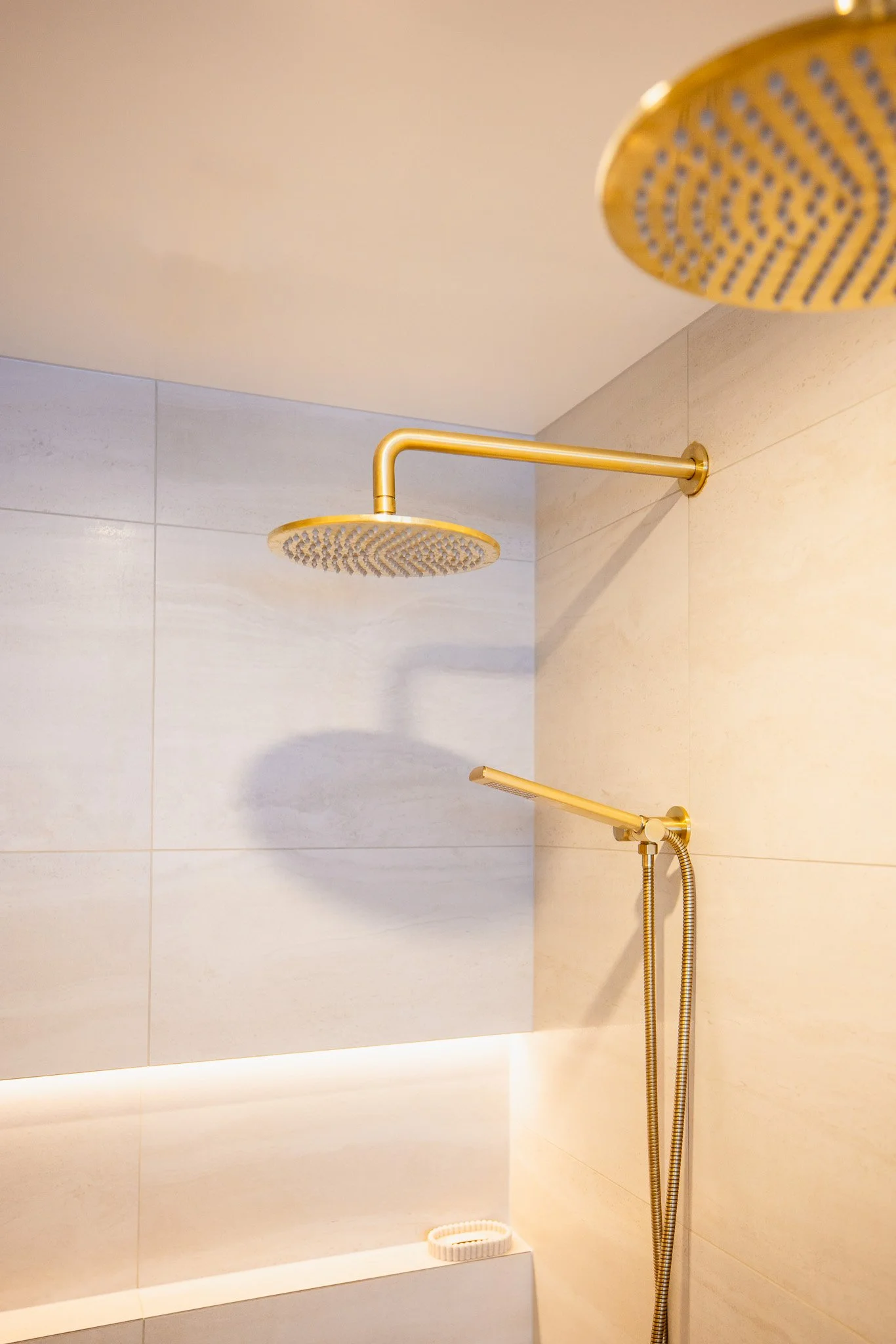 A modern shower with a large, round, gold-colored rainfall showerhead mounted on the ceiling and a handheld showerhead on a flexible hose attached to the wall, all in a light-colored tiled shower enclosure.