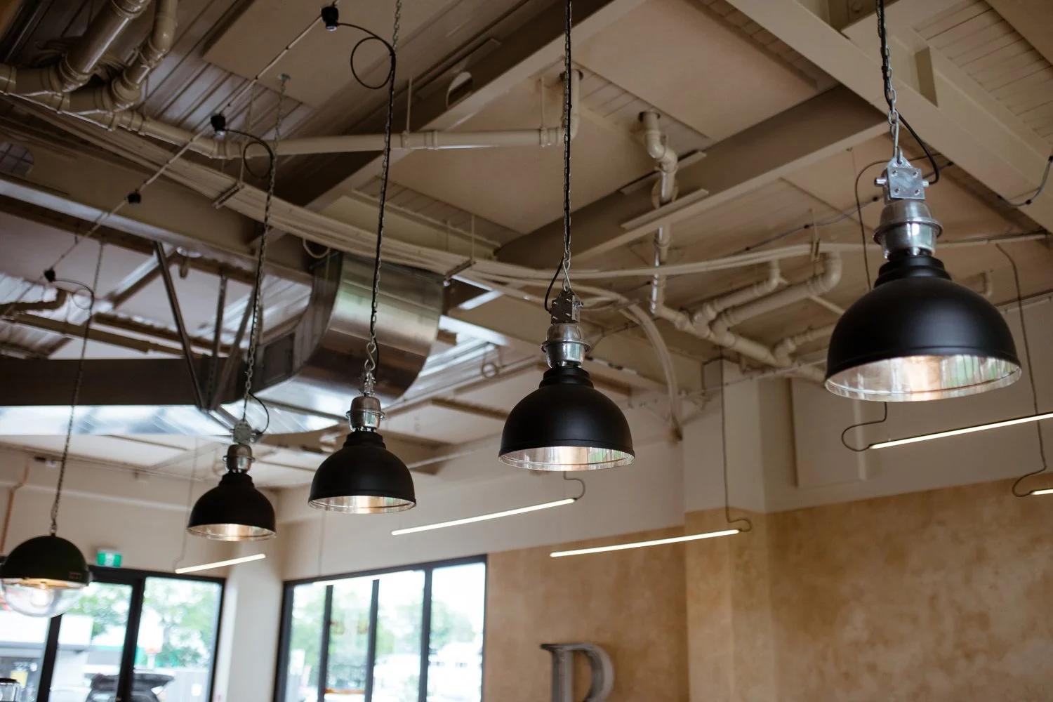 Interior view of a modern industrial-style space with black hanging pendant lights, exposed ceiling pipes, and large windows