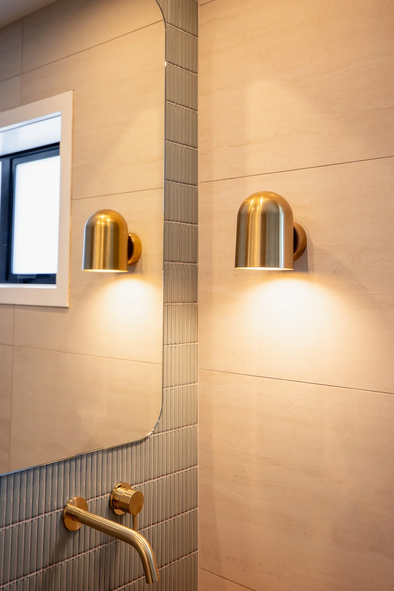 A bathroom corner with two brass wall-mounted lamps illuminating a beige wall and a small window, and a brass faucet below a decorative mirror.