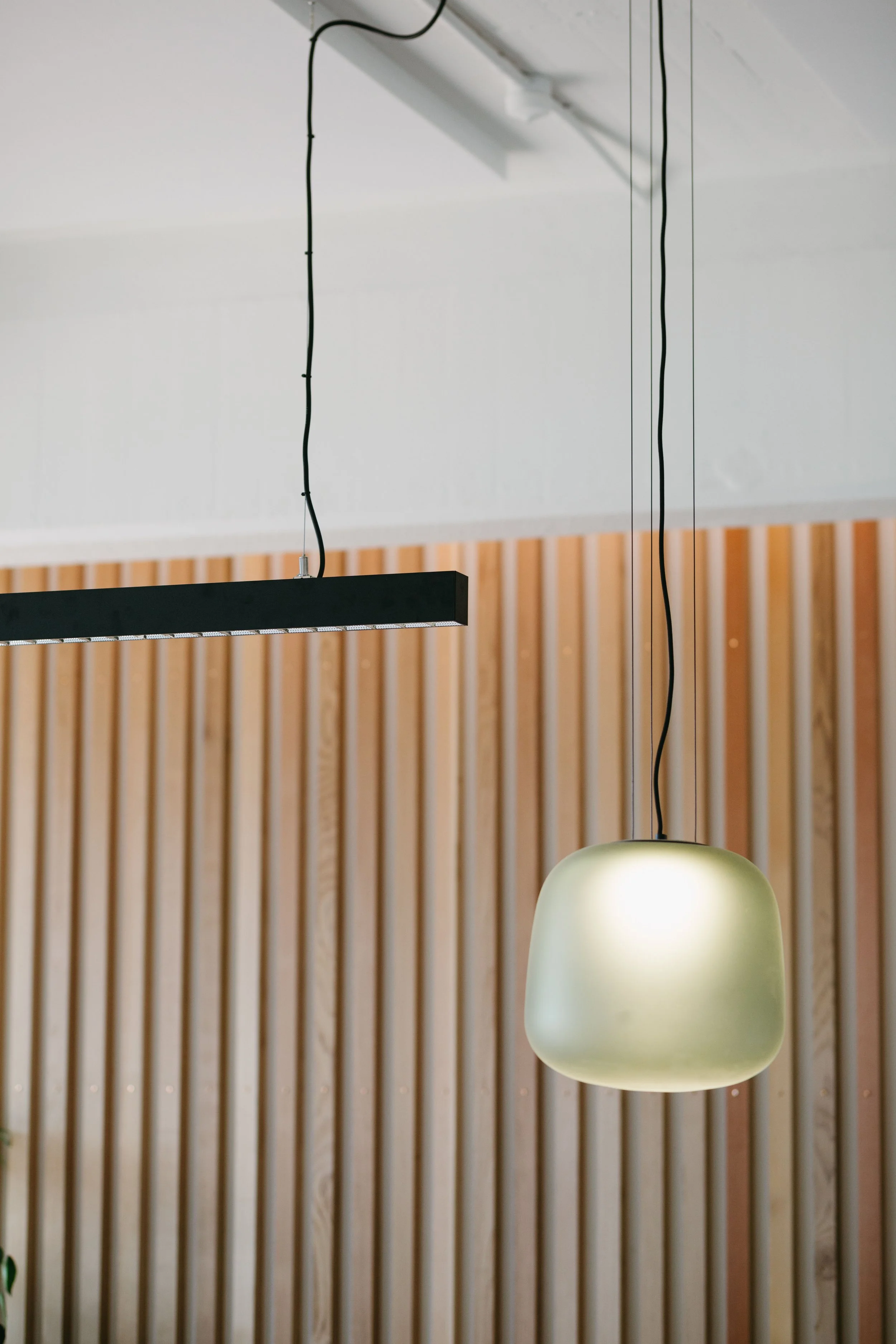 A modern hanging pendant lamp with a frosted glass shade, suspended from the ceiling with black cords, against a wooden slat wall.
