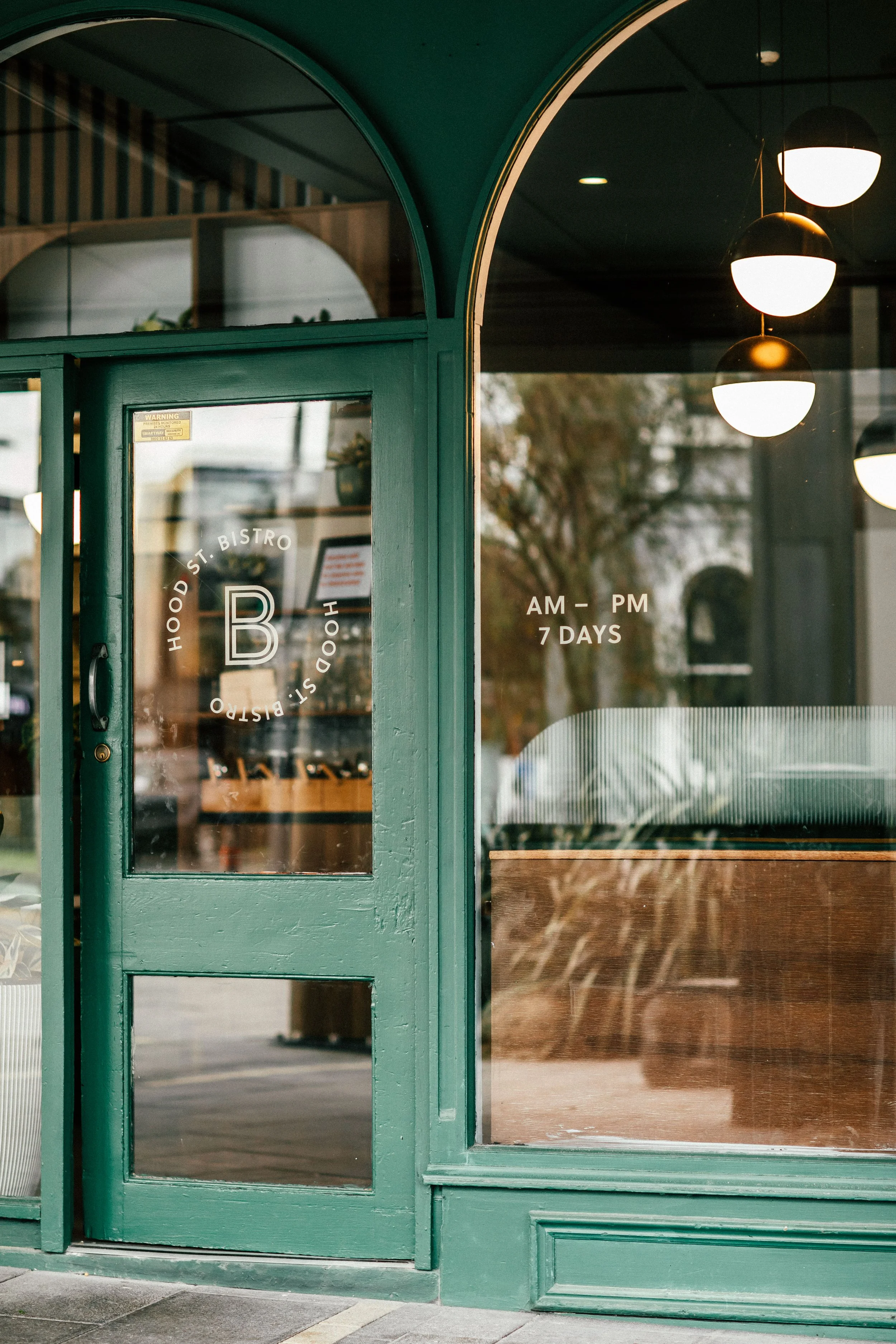 Green storefront with a glass door and window, displaying the name 'Hood St. Bistro' and hours 'AM - PM 7 DAYS'. Inside, round hanging pendant lights and seating are visible.