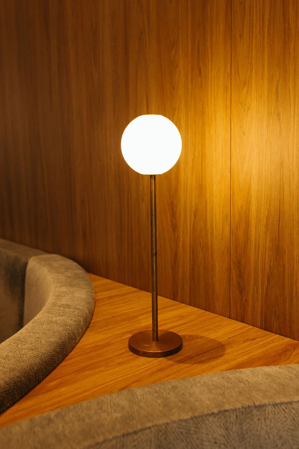 A table lamp with a round white shade and brass stem on a wooden surface, against a wooden wall background.