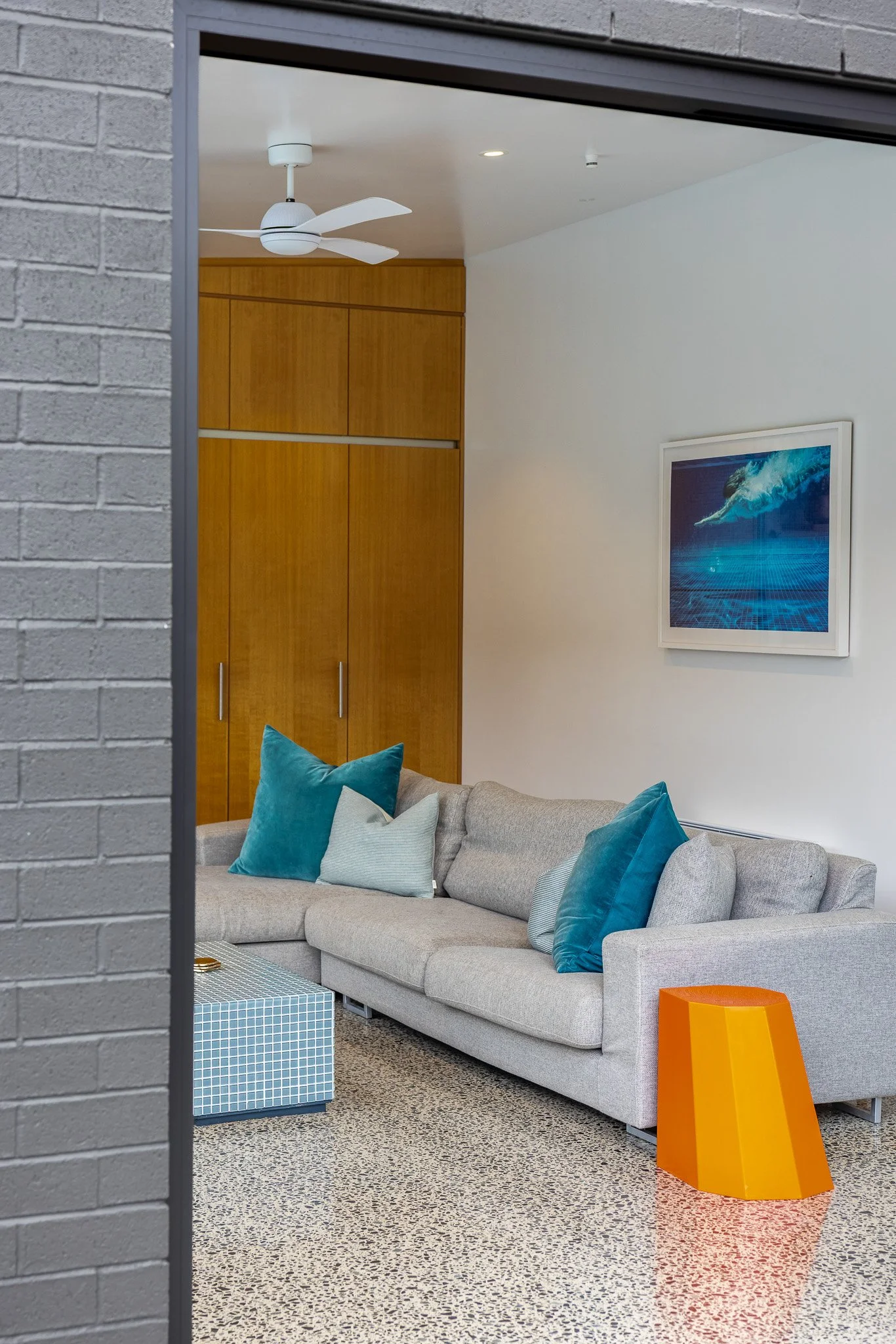View through an open sliding door into a contemporary living room with a gray sofa, blue and white pillows, a wooden closet, a white ceiling fan, a framed ocean-themed picture, and a bright orange side table.
