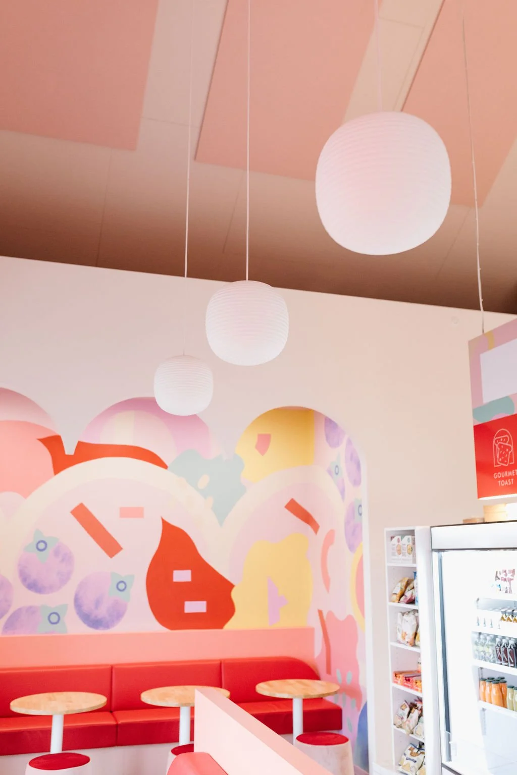 Interior of a pastel-colored cafe or snack shop with white hanging paper lanterns, a colorful abstract mural featuring blueberries, strawberries, and swirls, and a red cushioned seating area with round wooden tables.