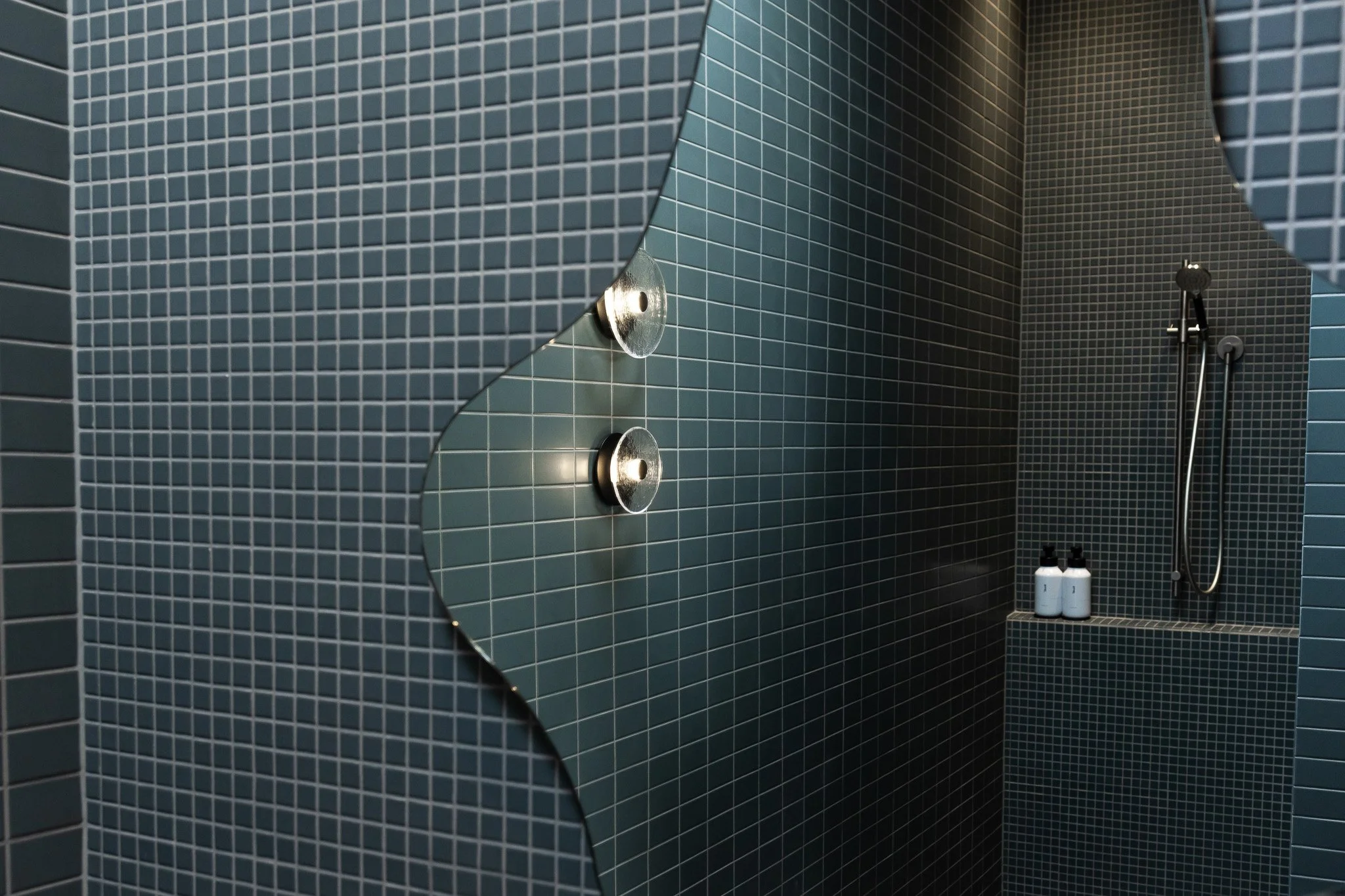 A modern shower area with blue tile walls, a handheld showerhead, and bottles of shampoo and body wash on a ledge.