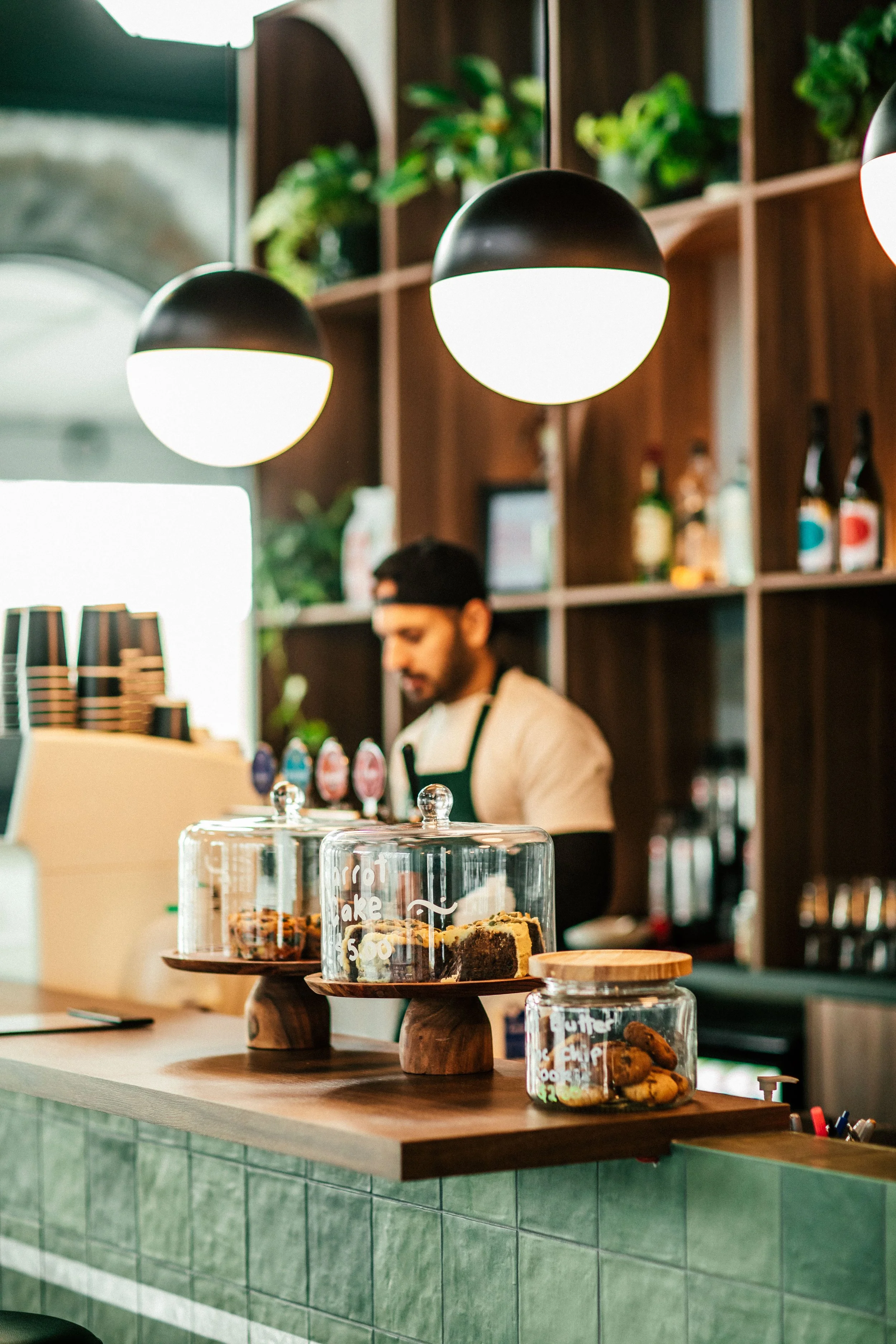 Counter with glass jars containing cookies and cake slices, a barista in the background at a coffee shop with dark wood shelves, plants, and hanging lights.