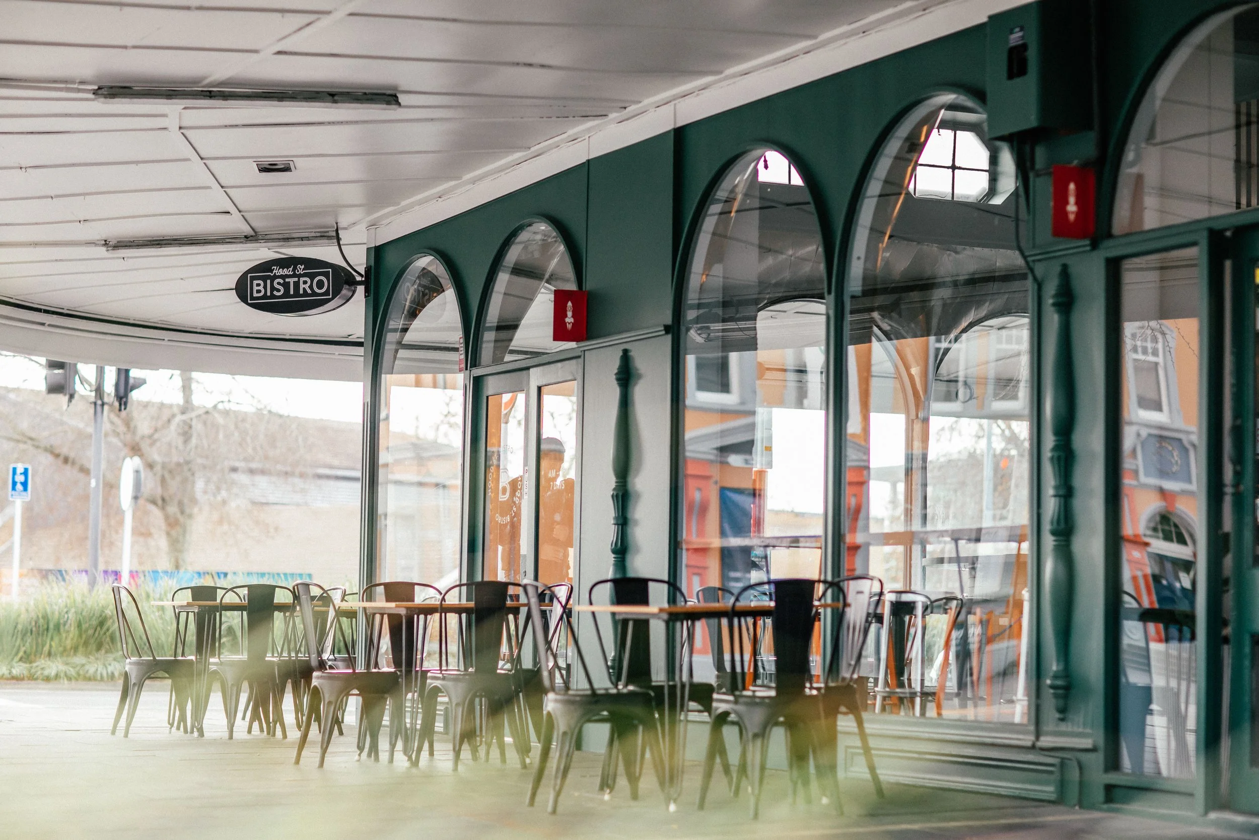 Empty outdoor cafe with colored chairs and tables under a covered patio, seen through large glass windows, with a sign hanging that says 'Hood St Bistro'.