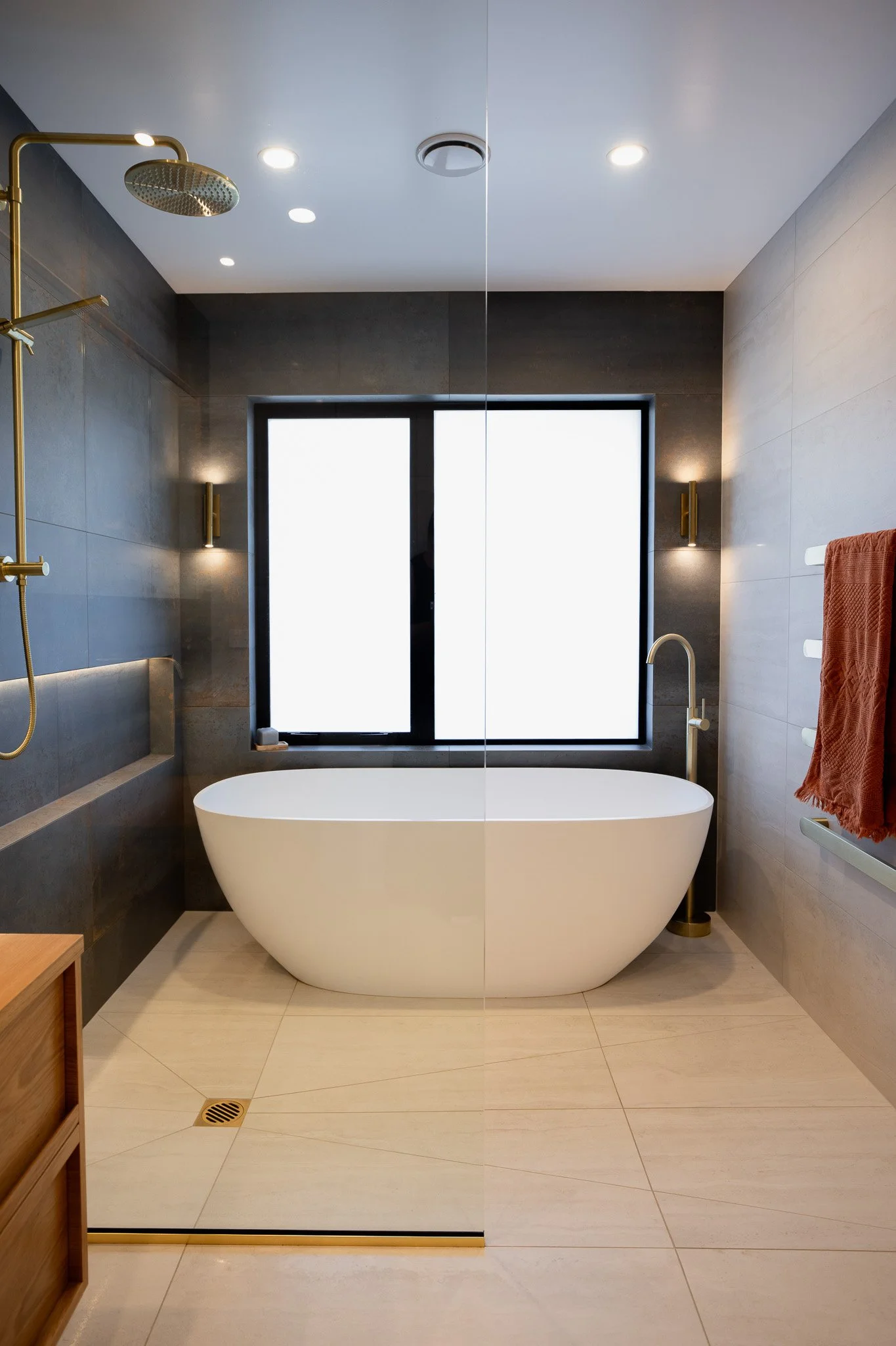 Modern bathroom with a free-standing bathtub in front of a large window, dark wall tiles, and a gold showerhead on the left.