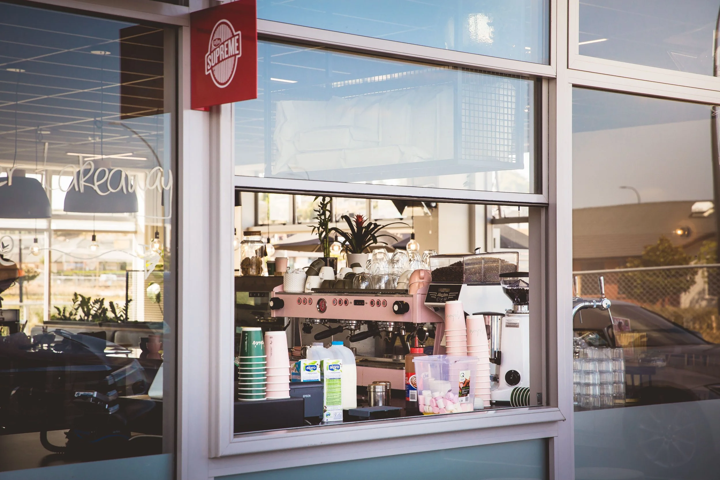 Coffee shop interior with a pink espresso machine, cups, a coffee grinder, and containers of ingredients seen through a large window.