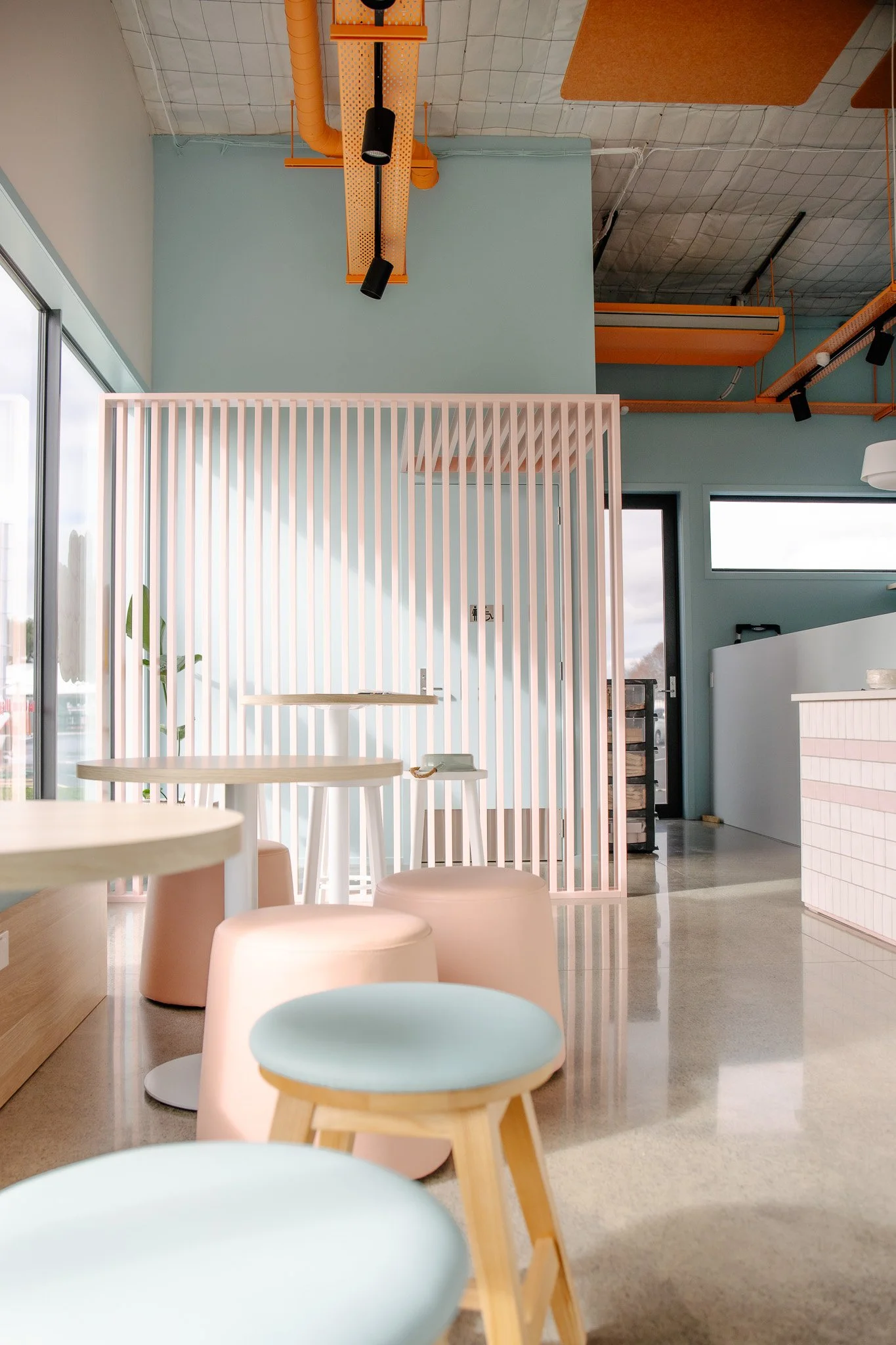 Interior of a modern cafe with pastel-colored furniture, light blue walls, and pink wooden slats creating a partition. There are round tables with stools, large windows allowing natural light, and ceiling fixtures in orange and black.