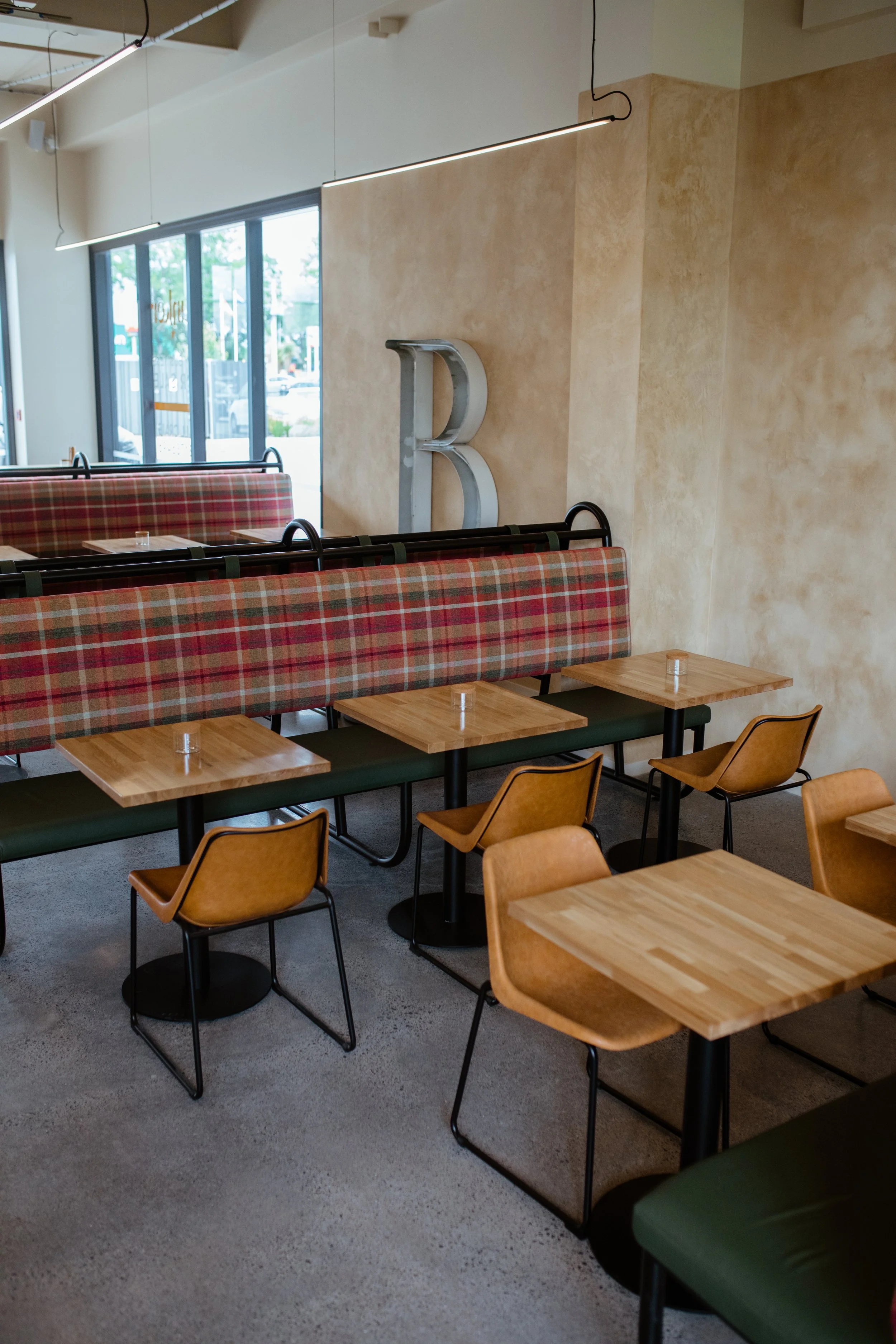 Empty restaurant with wooden tables, leather chairs, and a patterned booth. Large window with outside view, modern lighting, and a metal letter 'B' on the wall.