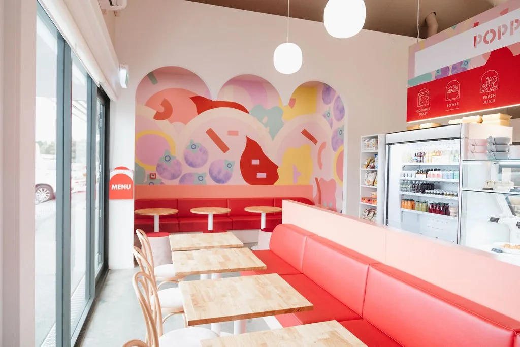 Inside a colorful ice cream shop with pink and white decor, featuring a mural with abstract shapes on the wall, red cushioned seating, wooden tables, and a refrigerator stocked with beverages.