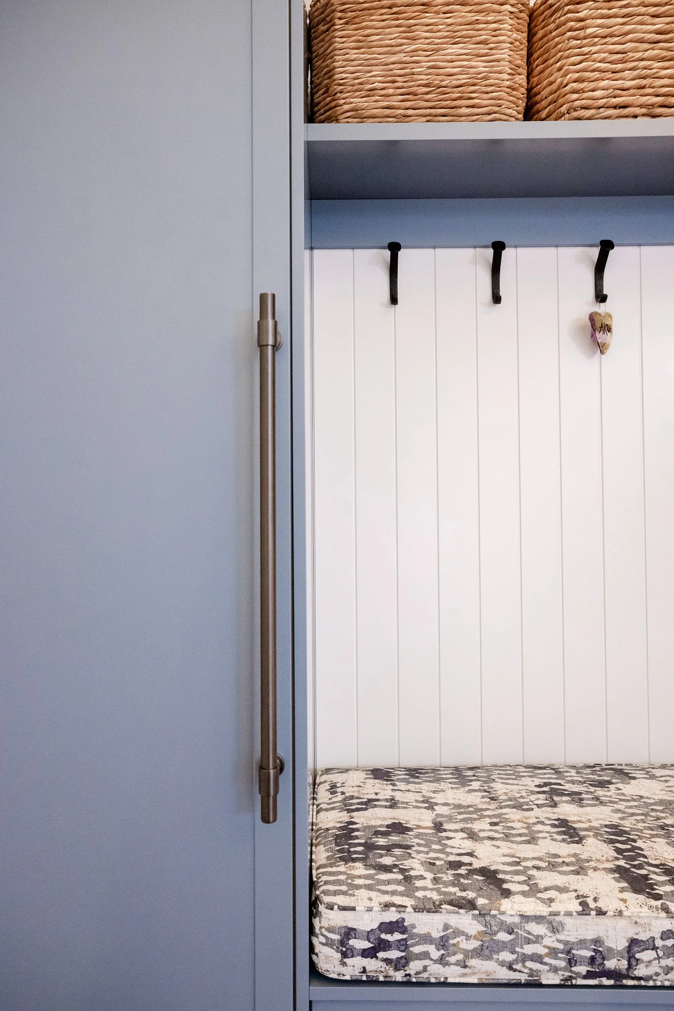 View of a closet with a light blue door on the left, a hanging rod with hooks and a heart-shaped decoration, a white-paneled wall, and a patterned mattress or cushion at the bottom.