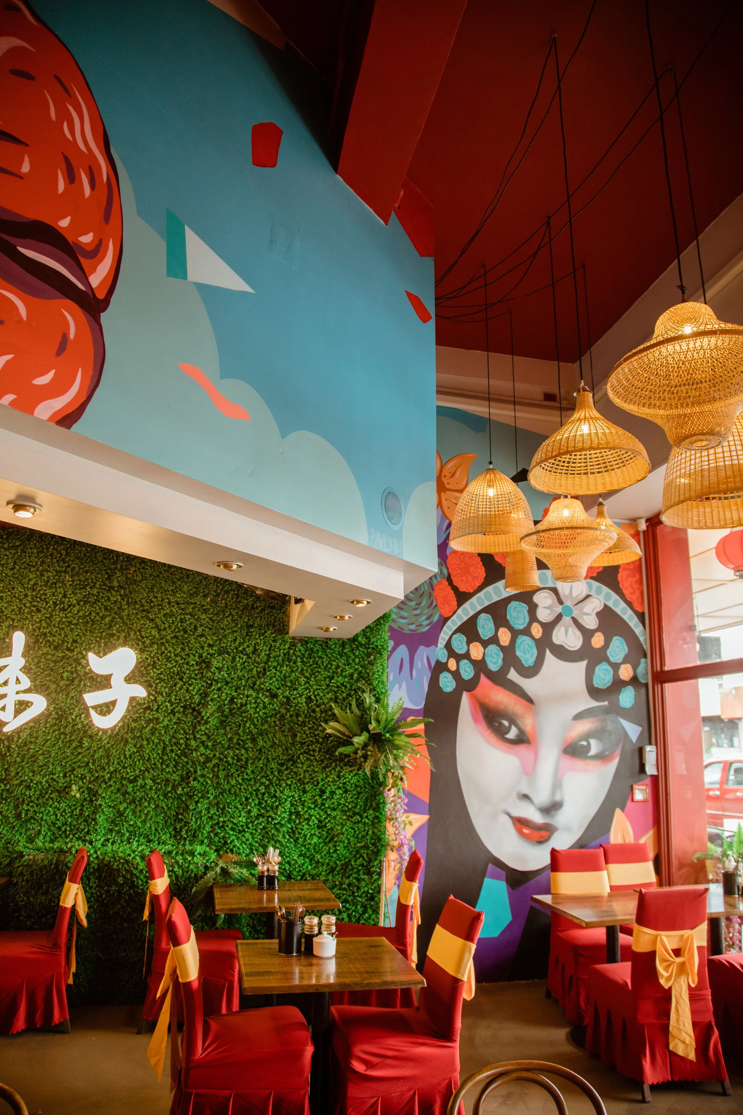 Colorful restaurant interior with modern Asian-style decor, including a large mural of a woman with traditional makeup, hanging wicker lamps, red chairs with yellow ribbons, and a green wall with Chinese characters.