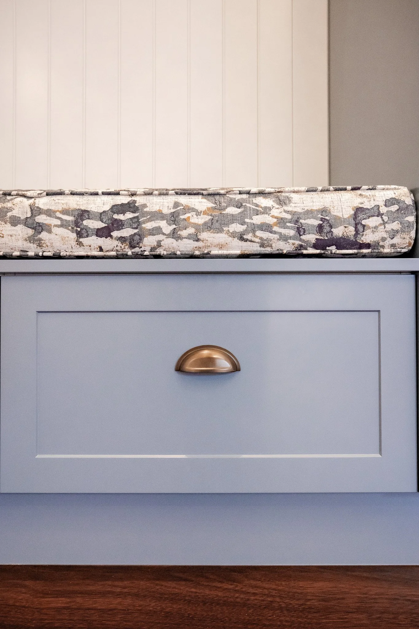 Close-up of a light blue kitchen cabinet drawer with a brass handle, a gray patterned countertop above, and a white wall in the background.