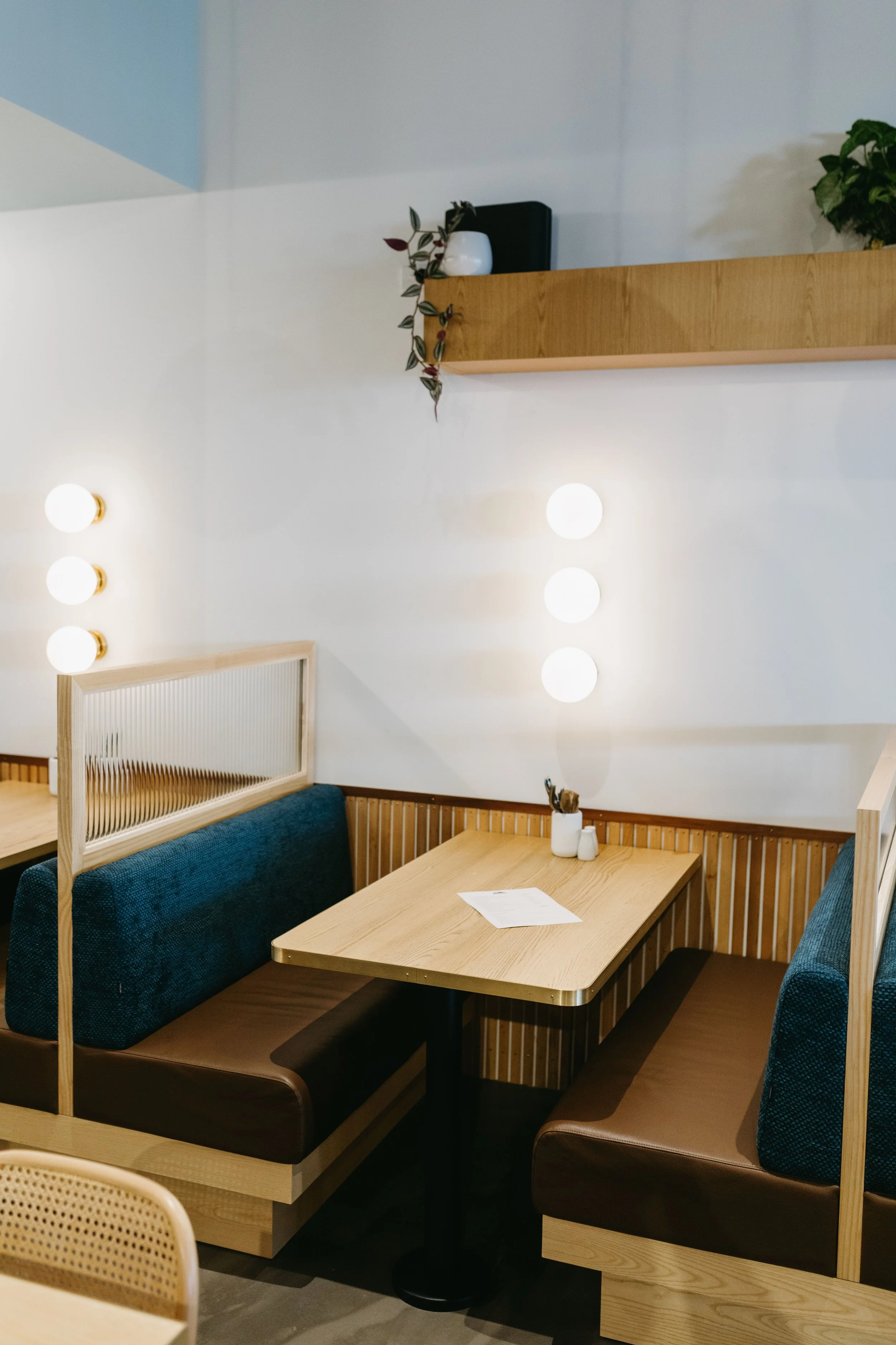 A cozy restaurant booth with a wooden table, brown and teal cushioned seating, and a white wall illuminated by four round wall lights. There is a paper and utensils on the table, with a shelf holding a plant, a white vase, and a black speaker above.