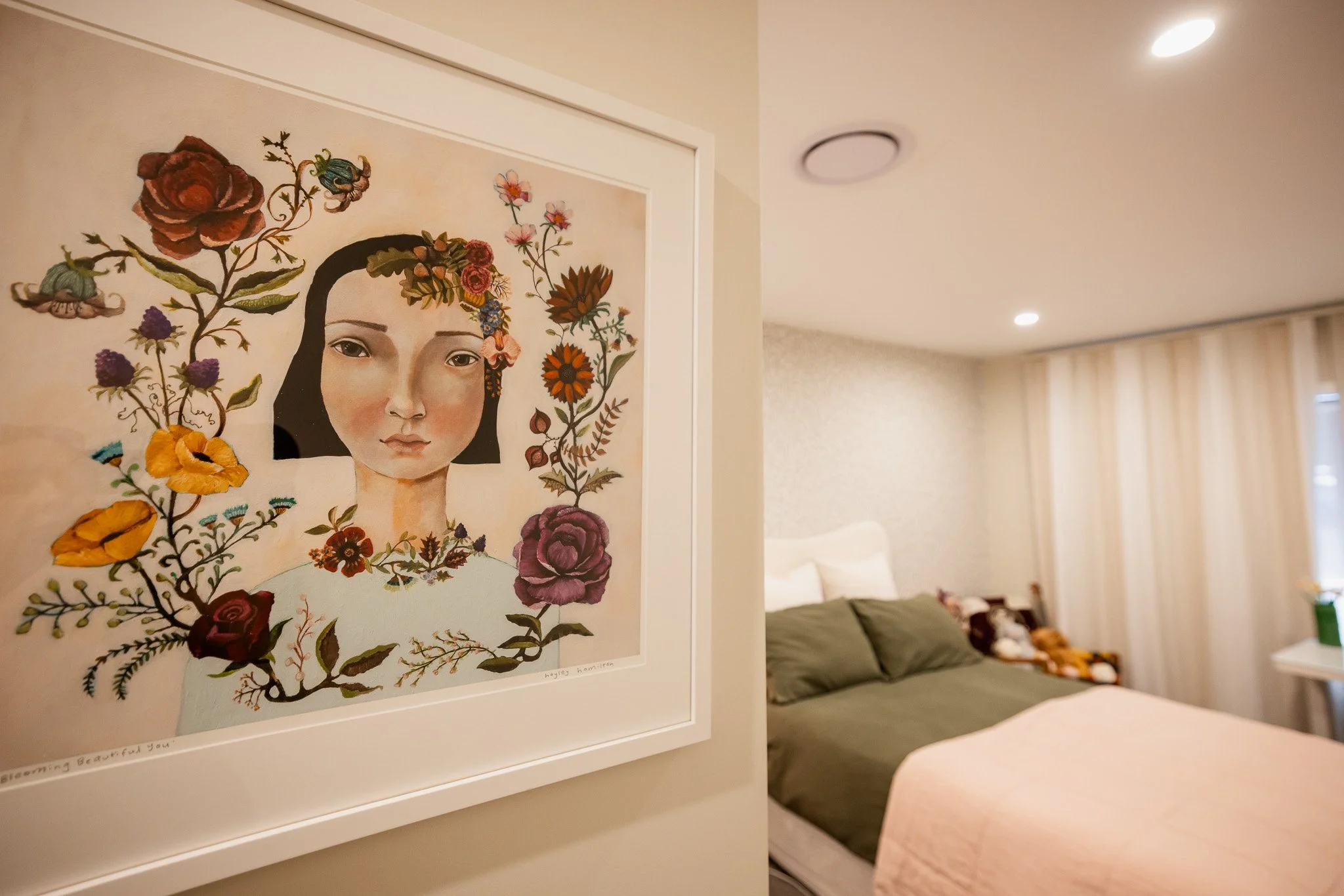 A framed artwork of a woman's face surrounded by colorful flowers hangs on a wall in a bedroom. The room features a bed with green and white bedding, a teddy bear, and a sitting area with curtains.
