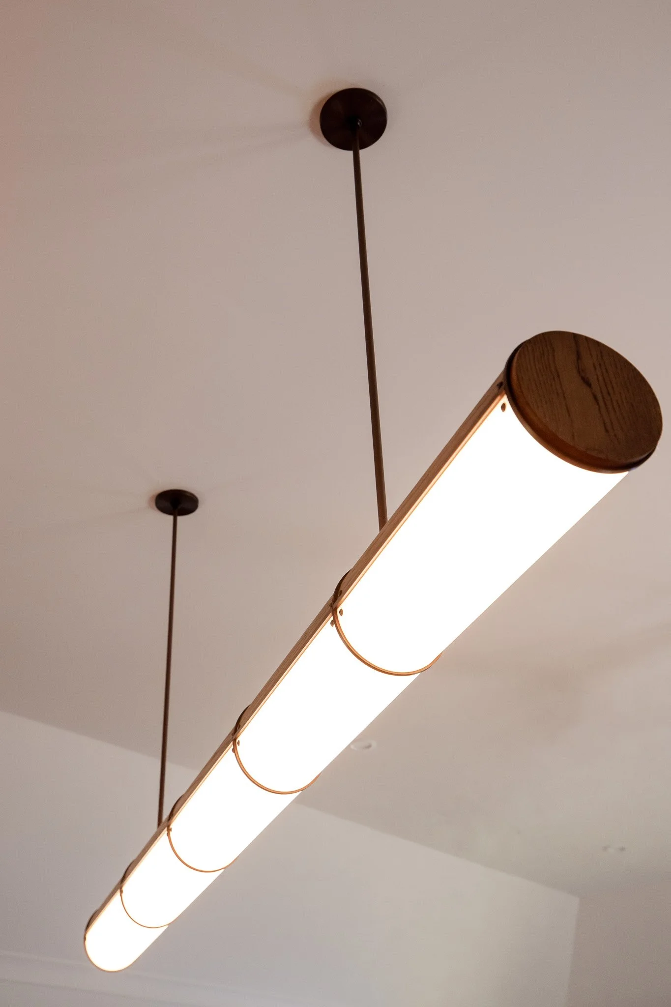 Modern ceiling light fixture with a long cylindrical white LED light, wooden end caps, and thin metal support rods, mounted on a white ceiling.