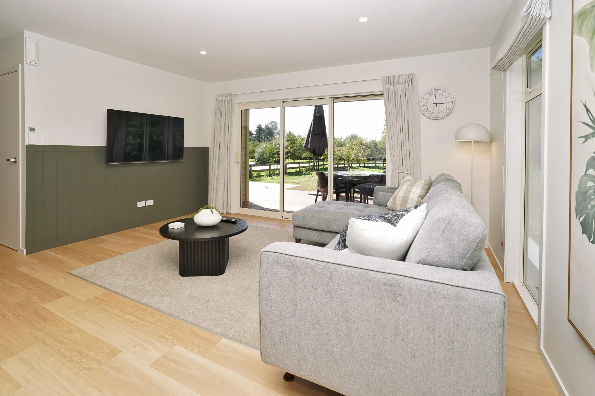 Bright living room with a gray sofa, beige carpet, black coffee table, wall-mounted TV, sliding glass door with curtains leading to patio, outdoor table with umbrella, and view of garden