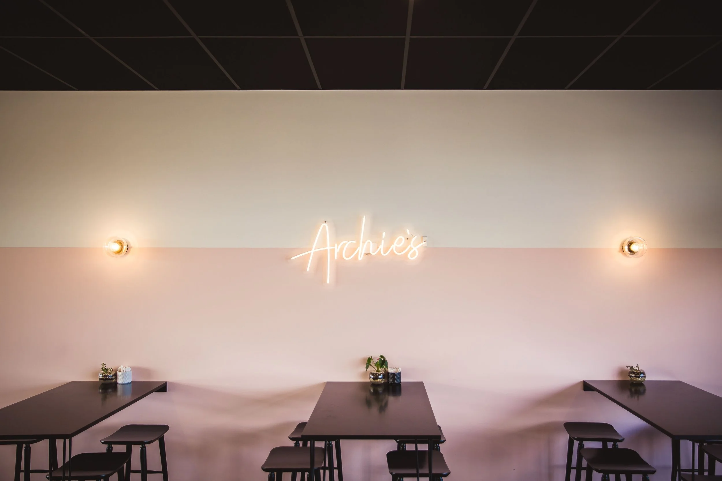 Interior of a cafe with three black tables, small potted plants, and a pink and white wall with a neon sign reading 'Arches'