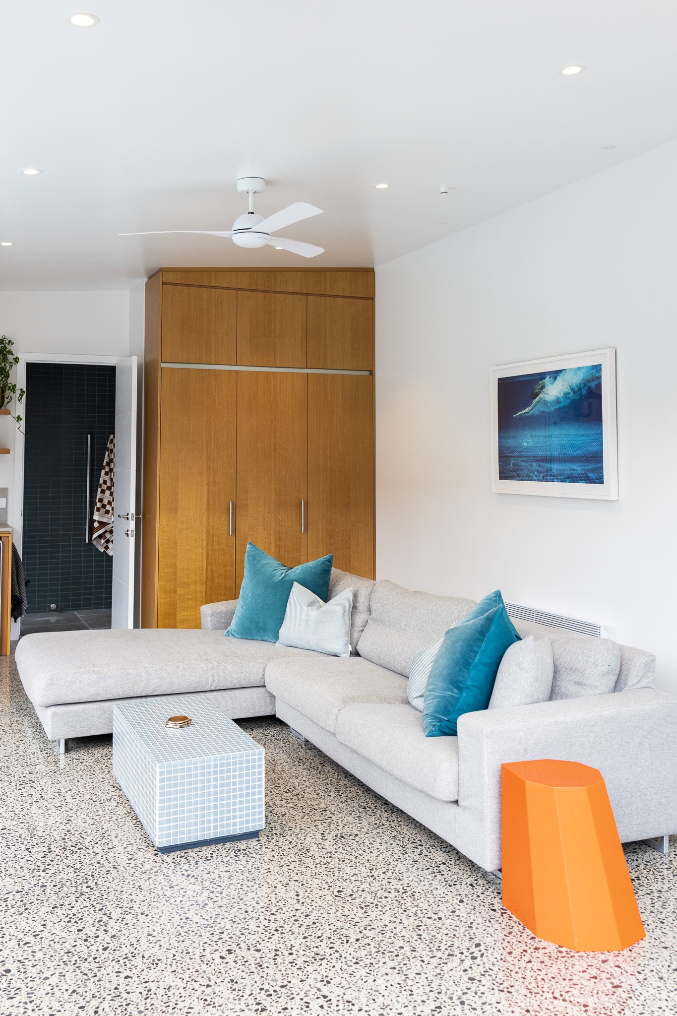 Modern living room with a light gray sectional sofa decorated with blue and white pillows, a white tiled coffee table, an orange side table, a wooden closet, framed ocean-themed picture, and a ceiling fan.