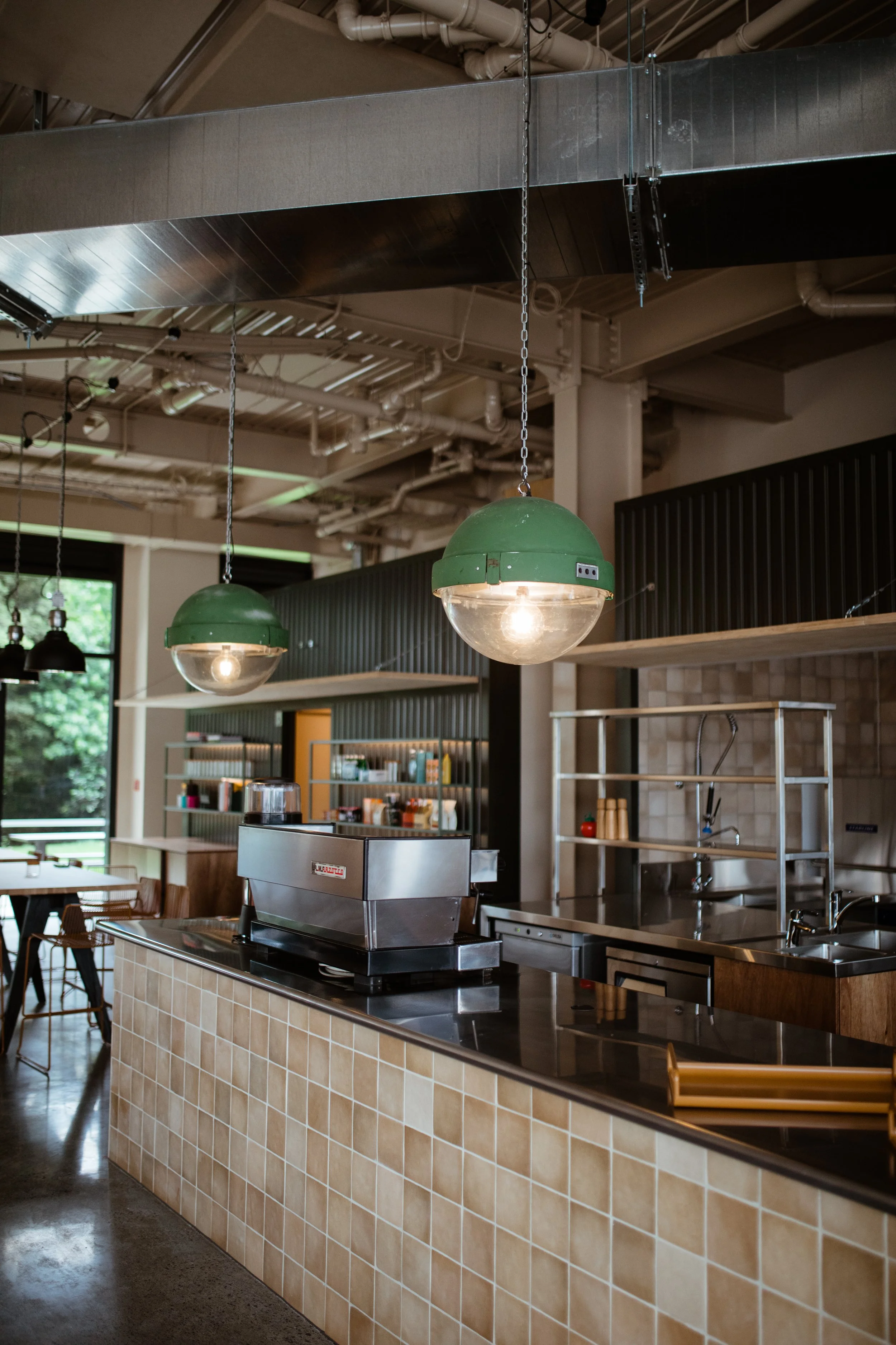Interior of a cafe or restaurant with a counter, hanging green pendant lights, shelves with condiments, and a coffee machine.