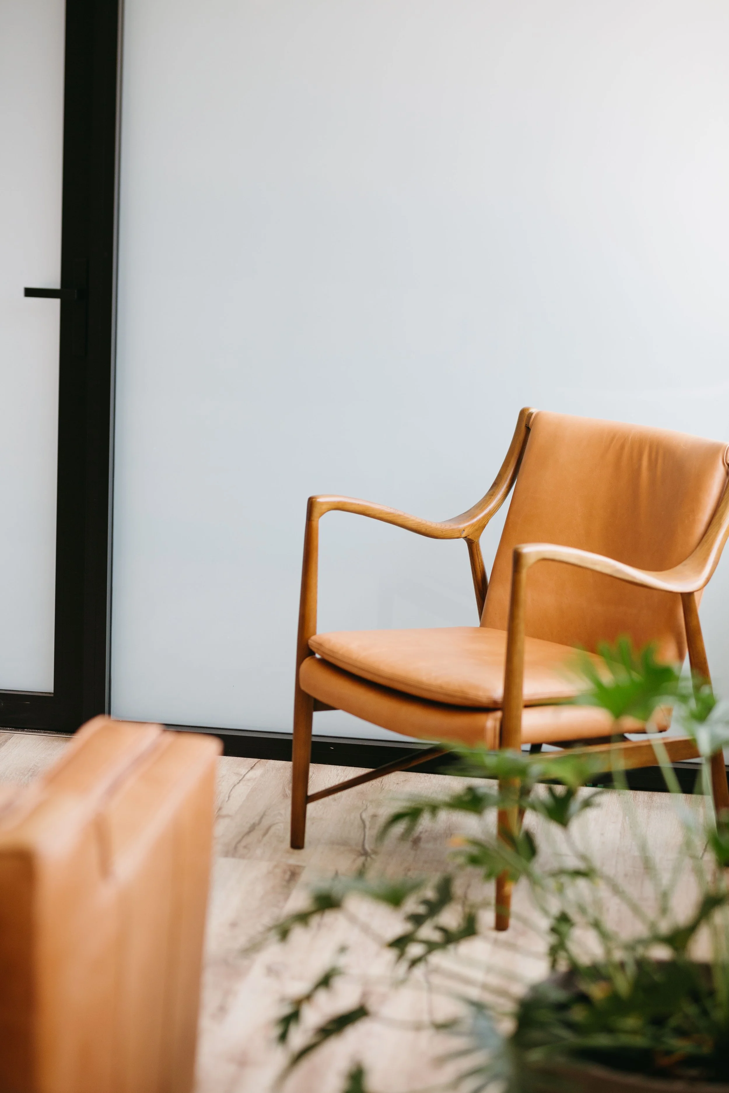 A cozy indoor scene with a wooden armchair with tan leather upholstery and a plant in the foreground, in a modern room with a wooden floor and a white wall.
