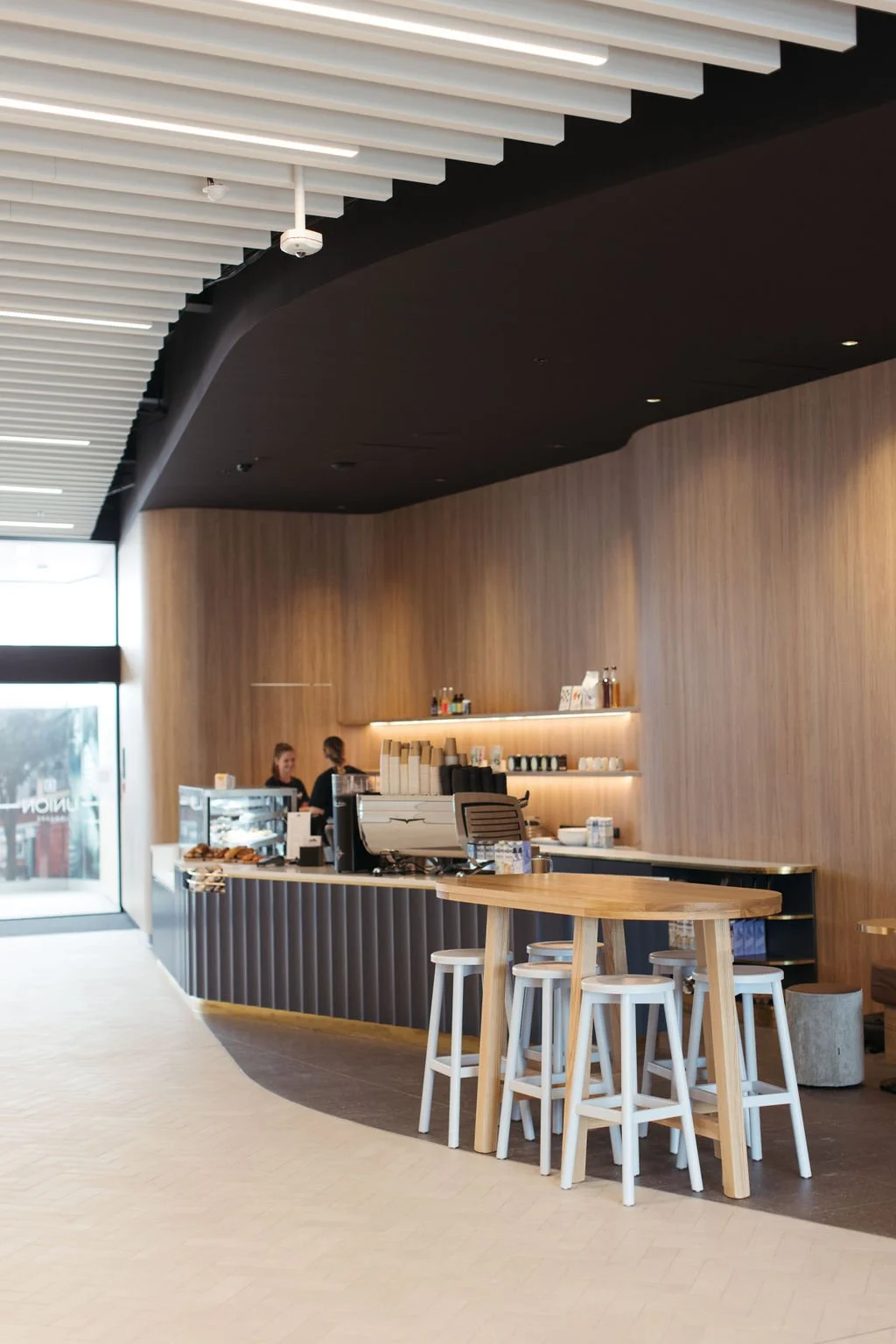 Modern coffee shop interior with a counter, barista, and seating area including stools and a high table. Wooden walls and ceiling with linear lighting.